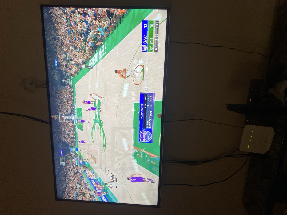 The image captures a television screen displaying a basketball video game, likely set indoors in a dimly lit room. The game features the Sacramento Kings playing against the Milwaukee Bucks.
On the top right of the screen, the scoreboard indicates "SAC" (Sacramento) has 13 points and "MIL" (Milwaukee) has 10 points. The game is in the "1st" quarter with "2:05" minutes remaining, and a number "23" is also visible, possibly indicating a shot clock. A digital player, identified as "G. Antetokounmpo" (Giannis Antetokounmpo), is visible on the court, highlighted by a ring around him. On the bottom left of the screen, player statistics for the "SACRAMENTO KINGS" are shown: "D. FOX" has 9 "PTS" and 4/4 "FES", while the "REST OF TEAM" has 4 "PTS" and 2/5 "FES". The virtual basketball court features the Milwaukee Bucks' branding, including "FEAR THE DEER".
To the right of the television, a white internet router or modem with several green indicator lights is visible. Various black cables and wires run behind the TV and connect to the router and other dark electronic devices situated below it. The ambient lighting of the room is dim, suggesting it might be evening or a low-light environment. No specific visual cues within the image confirm the provided location context of Lakka, Sierra Leone.