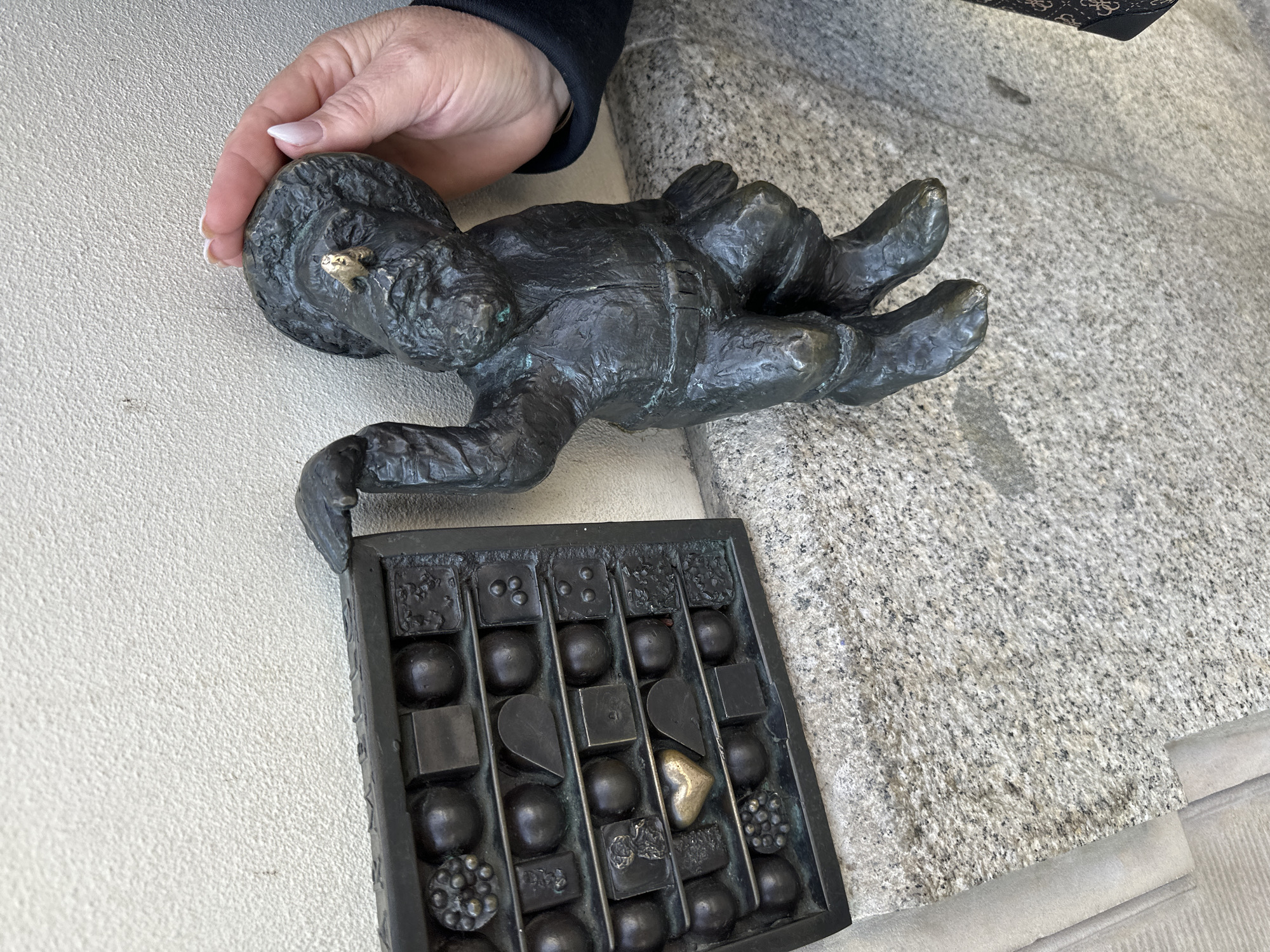A close-up shot shows a bronze dwarf statue and a box of chocolates. The dwarf is depicted lying on his stomach, with his right hand being touched by a human hand. The dwarf has a golden element on its head. Below the dwarf lies a bronze box of chocolates with various shapes and textures, including a golden heart-shaped chocolate. The scene is set on a rough stone surface, with a smoother surface visible on the left. This is a statue of a "Wrocław dwarf", specifically "Słodyczek" (sweet tooth), one of the many dwarf statues spread around the city of Wrocław, Poland. The statue represents a sweet tooth and is often placed near candy stores or confectioneries.