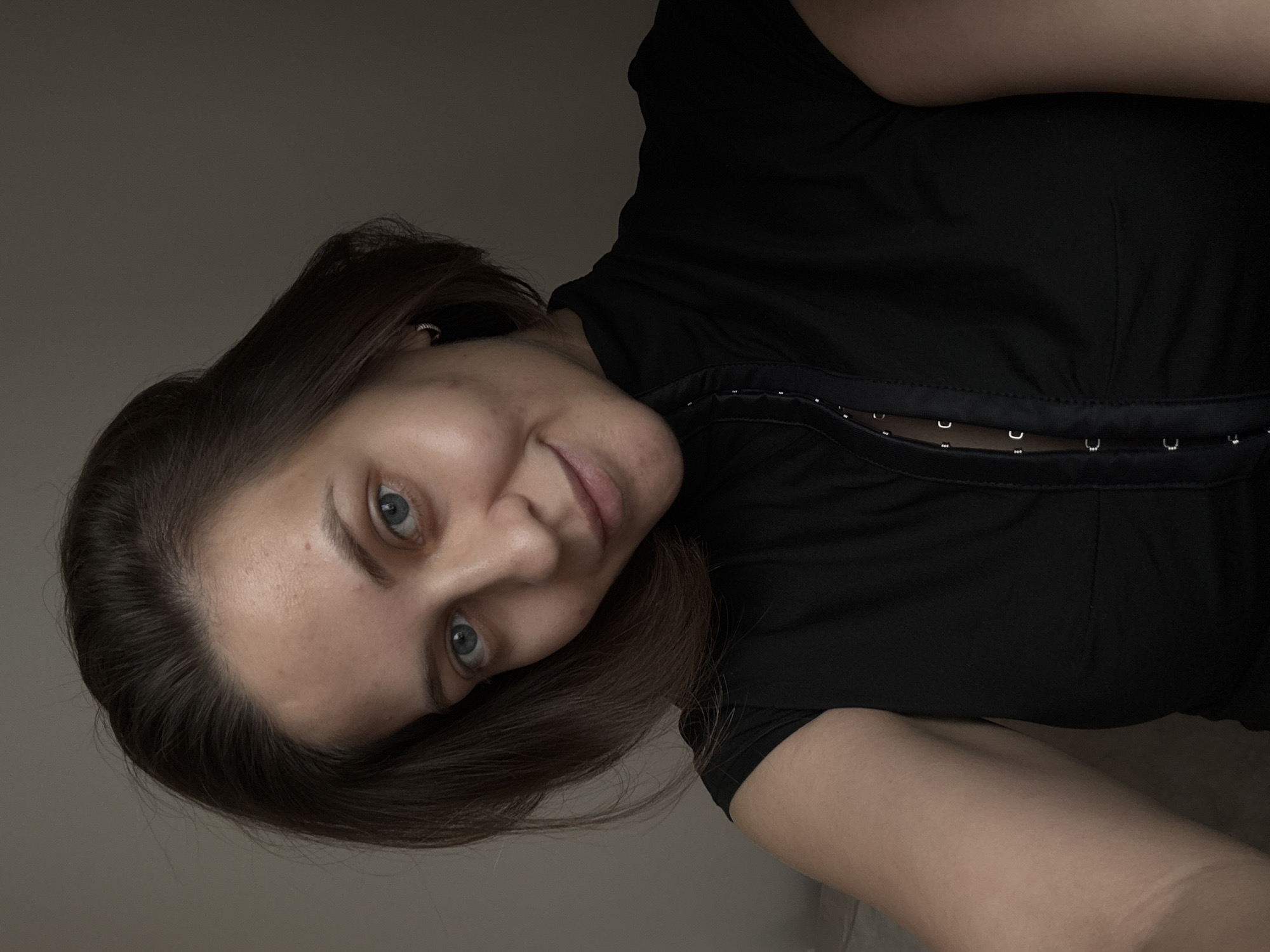 A medium-shot photograph shows a woman with brown hair and blue eyes. She is looking at the camera and smiling. She is wearing a black top. The background is a plain wall. The lighting is dim, with shadows obscuring parts of the image. The woman's skin appears clear with a subtle glow. The overall impression is a casual, self-taken portrait. There is no discernible information about the location within the city of Pervomaysky, Russia, nor any environmental details that would indicate this.