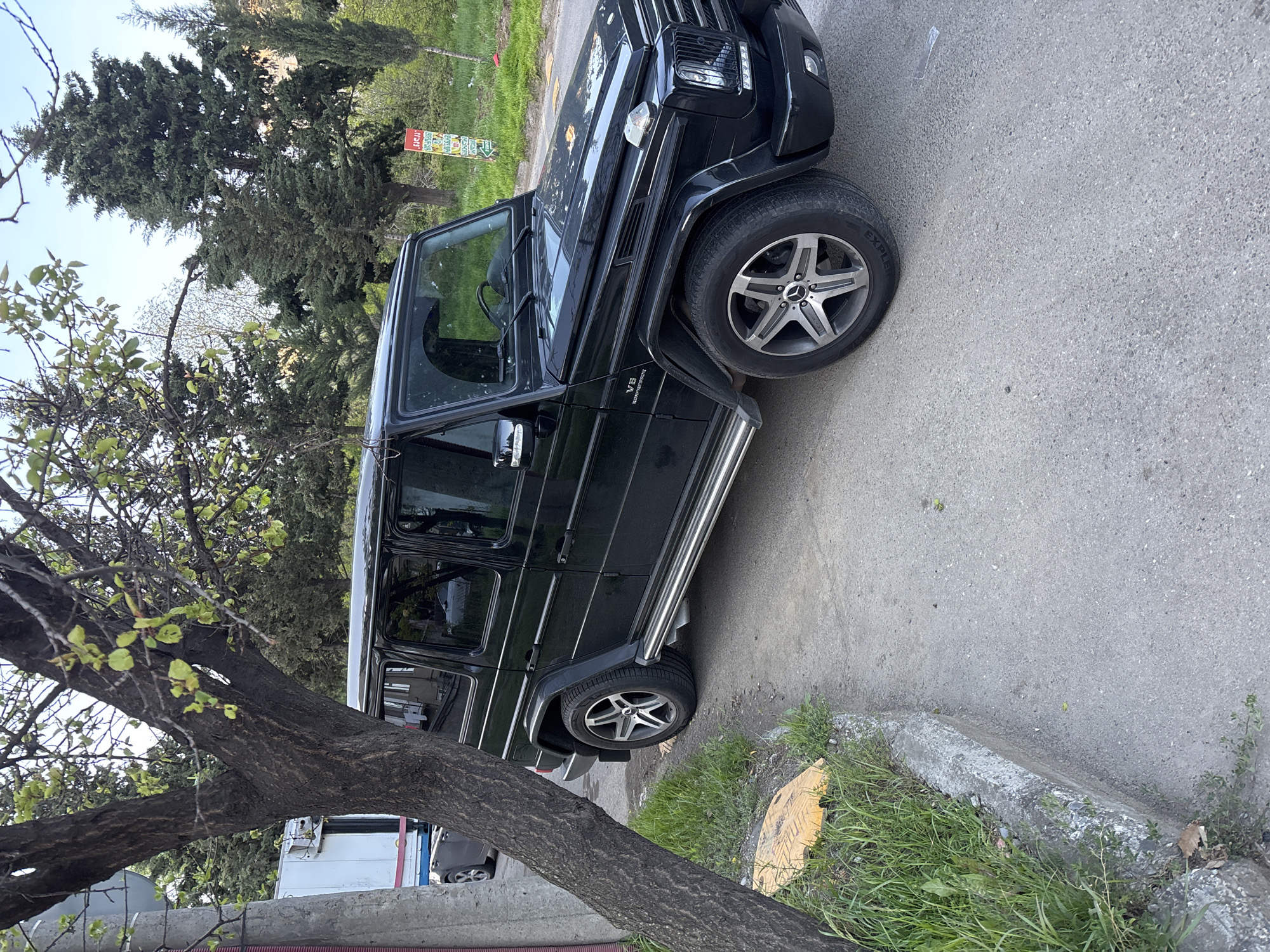 A black Mercedes-Benz G-Class SUV is parked on the side of a street in Tbilisi, Georgia. The car is positioned at an angle to the curb, partially blocking the sidewalk. The vehicle is facing up and to the right.
The setting is an urban area with trees and greenery visible. A tree trunk is prominent in the foreground to the lower left, partially obscuring a parked white vehicle further back. The asphalt road has some visible imperfections. There is grass growing along the curb.
On the car, a "V8 Kompressor" badge is visible on the side. The wheels have a five-spoke design with the Mercedes-Benz logo in the center. The car is equipped with side steps. A sign is visible in the background with orange and green coloring.