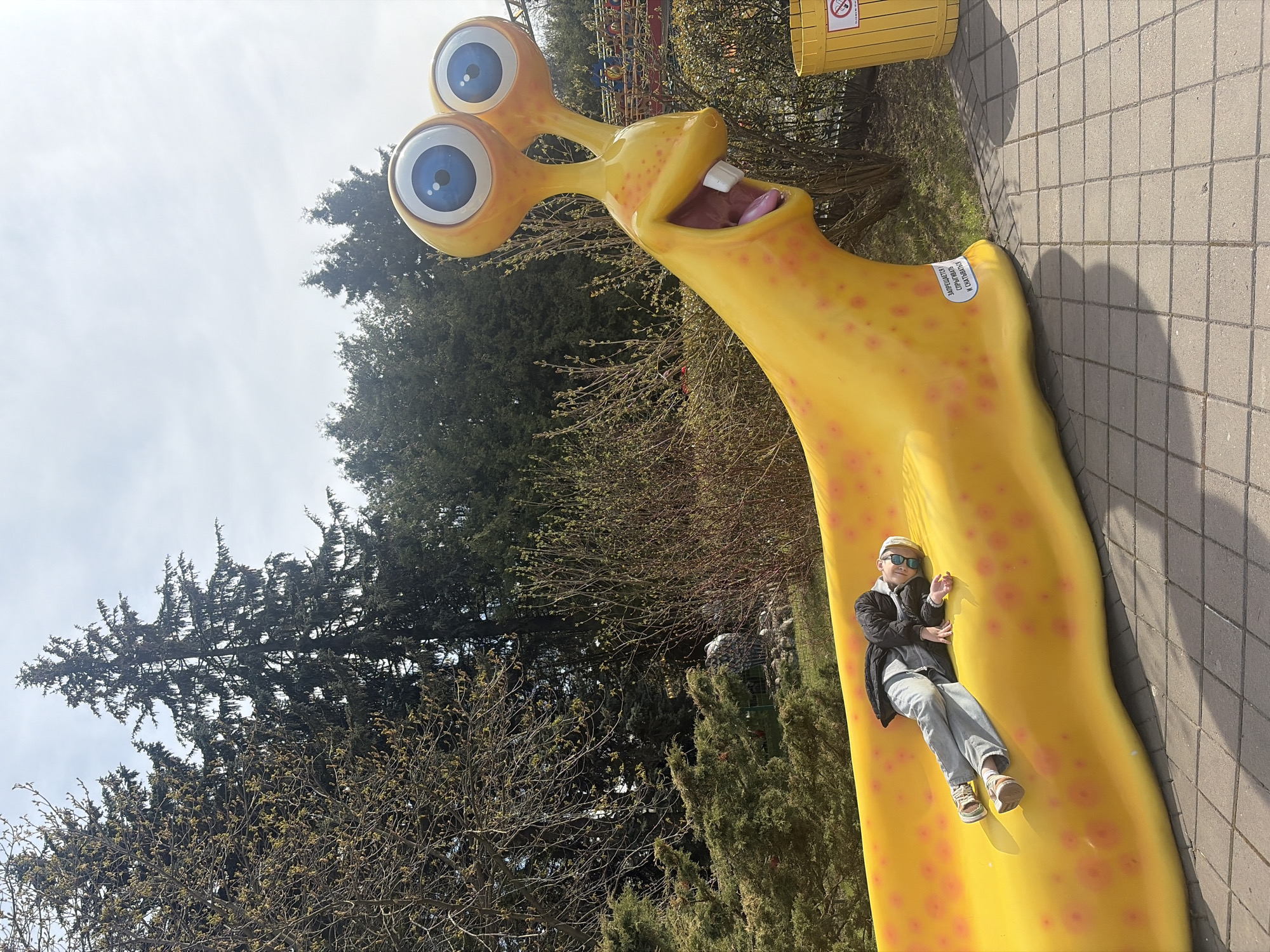 This outdoor image, taken in Saint Petersburg, Russia, features a large, bright yellow, slug-like sculpture with a person lying on it. The sculpture has a cartoonish appearance, with two prominent blue, googly eyes on stalks, a wide-open mouth revealing white teeth and a pink tongue, and numerous reddish-orange spots adorning its body.
A child or young adult is comfortably reclined on the sculpture's curved surface. They are dressed in a dark jacket, light-colored jeans, a beige baseball cap, and sunglasses, with their left hand resting on the sculpture and their legs stretched out. The person appears relaxed, posing for the camera.
The scene takes place on a bright, sunny day, evident from the distinct shadows cast on the light gray paved ground, which is composed of square tiles. In the background, a variety of trees are visible, including both deciduous trees with early spring buds and dark evergreen trees, suggesting a park or recreational area. A clear blue sky is visible above the trees. A yellow trash can with a "no smoking" symbol is partially visible in the upper right background.
A white sticker with Cyrillic text is affixed to the sculpture near the person's right hand. The text reads: "ЗАПРЕЩАЕТСЯ! СТРОГО СТОЯТЬ И ОБЛОКАЧИВАТЬСЯ!", which translates to "FORBIDDEN! STRICTLY STAND AND LEAN!".