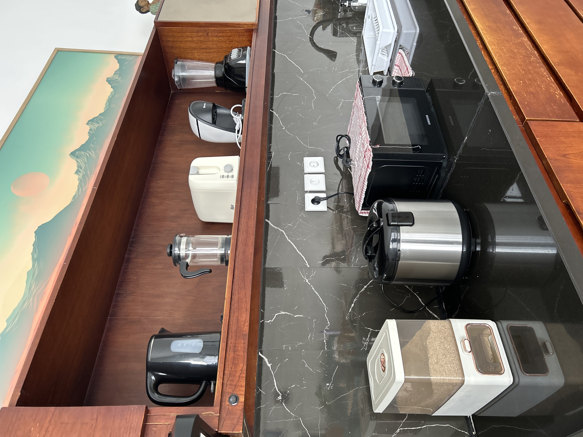 This is a medium-shot, eye-level interior photograph of a modern kitchen counter in Tembuku, Indonesia. The scene is dominated by dark, polished granite countertops and warm, wooden cabinetry. On the upper shelf, a collection of kitchen appliances is neatly arranged: a black electric kettle, a glass French press, a white toaster, a compact coffee maker, and a black blender. Above the appliances, a large framed artwork depicting a pastel-colored mountain range under a setting sun is displayed on the wooden wall.
On the countertop below, a white cereal dispenser filled with rice stands to the left. Next to it is a silver-colored rice cooker. Further to the right is a black Samsung microwave oven covered with a red and white checkered cloth. A white dish rack sits to the right of the microwave, with a faucet visible behind it. Three white electrical outlets are mounted on the dark tiled backsplash behind the microwave. The overall impression is one of a clean, well-organized, and functional kitchen space.