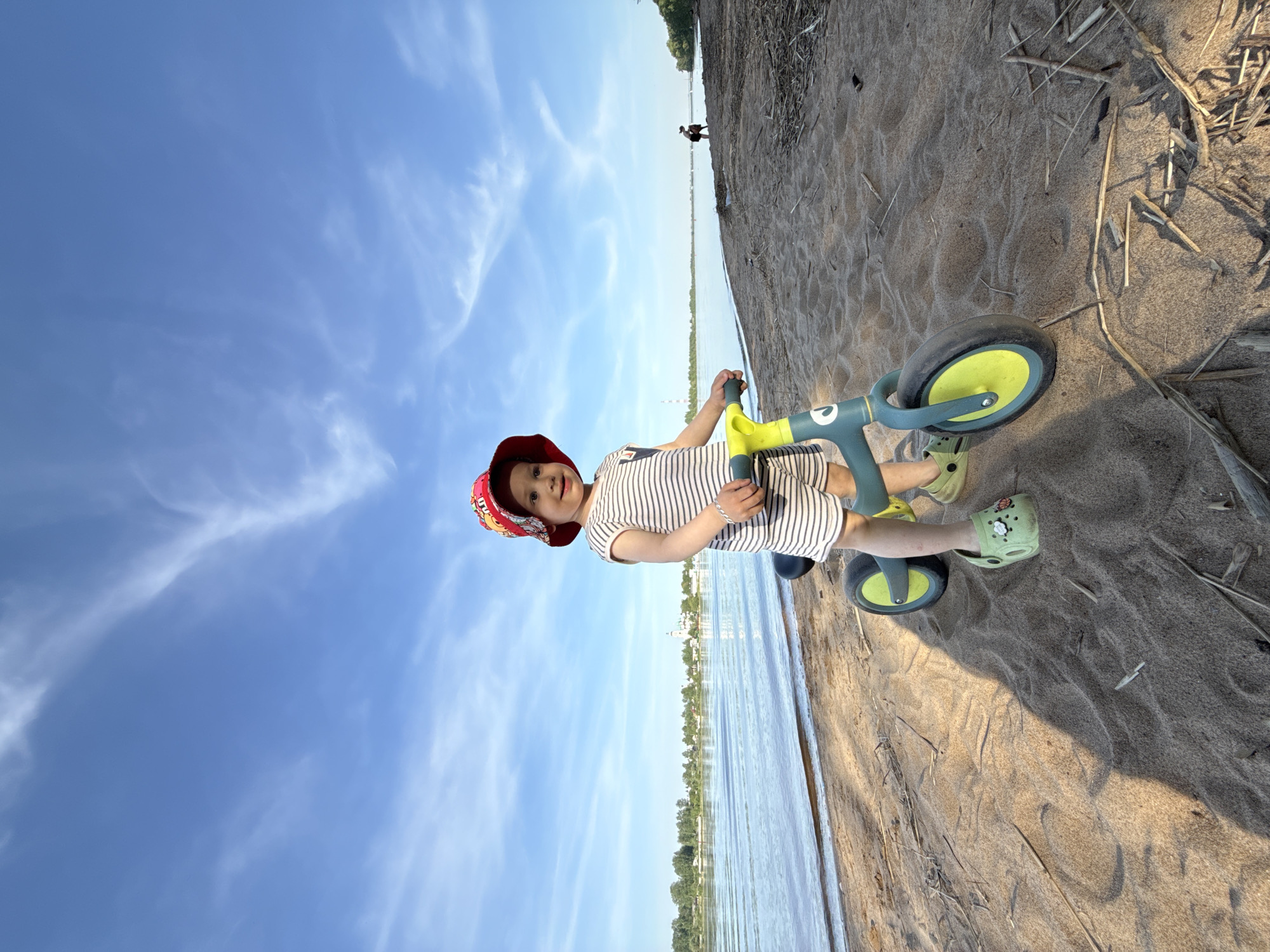 A young child in a striped outfit and a red sun hat stands on a balance bike on a sandy beach in Шебунино, Russia. The sky is blue with scattered clouds, and a body of water stretches along the horizon, separating the beach from trees on the opposite shore. A person walks on the beach in the distance. The child wears green shoes and holds the handlebars of the balance bike. Sticks and debris litter the sand around the bike.