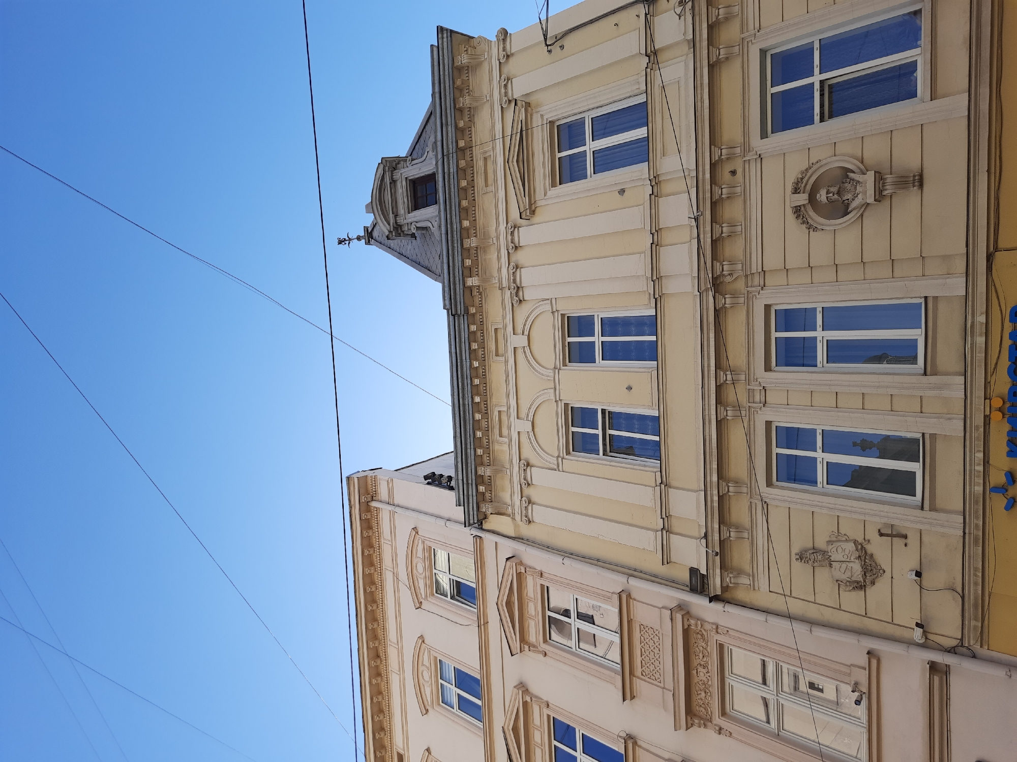 The image depicts the facades of two adjacent buildings in Lviv, Ukraine, viewed from a low angle looking upwards towards a clear blue sky. The buildings exhibit classical architectural styles with ornate detailing. The building on the left is painted a pale pink and features a series of evenly spaced windows with white frames and blue curtains. Decorative elements above some windows include triangular pediments and intricate stucco work below. The building on the right is painted a pale yellow and has a more complex facade. It features vertical fluted pilasters, decorative cornices, and arched window surrounds. One prominent feature on this building is a stucco relief of a bust within a circular frame, positioned between two windows. The roofline of the yellow building culminates in a mansard roof with dormer windows and a decorative finial. Electrical wires crisscross the sky above the buildings. The lower portion of the yellow building reveals a yellow storefront with blue lettering. The overall scene suggests an urban setting with historical architecture.