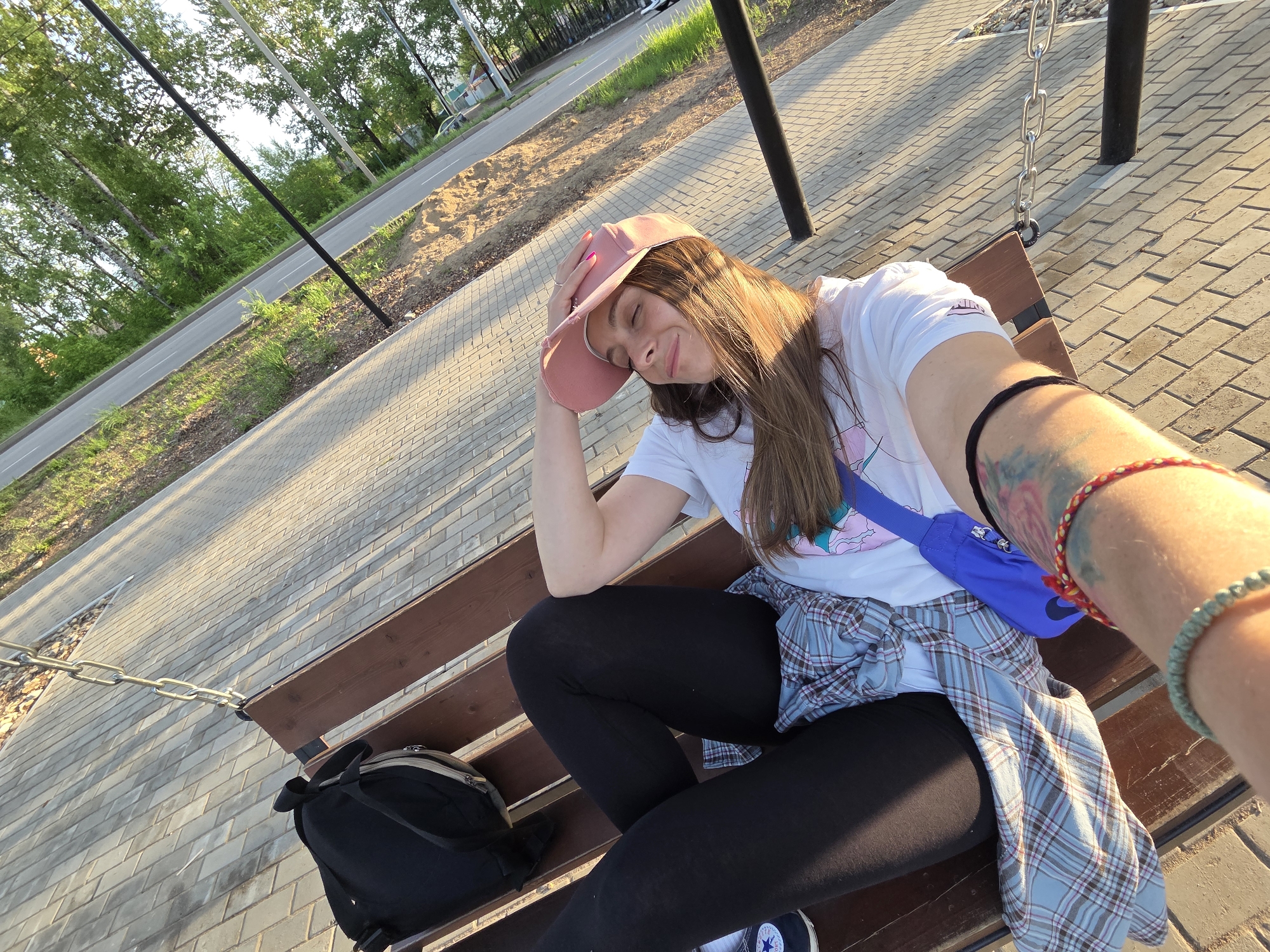A young woman sits on a park bench taking a selfie. She wears black leggings, a white t-shirt, a pink cap, and a blue fanny pack. A plaid shirt is tied around her waist. A backpack sits beside her on the bench. Her left arm rests on her head, and her right arm is extended towards the camera, revealing tattoos and bracelets. The bench is part of a swing set, with metal chains visible. The ground is paved with rectangular bricks. A road and trees are in the background. Based on visual clues and provided information, the scene likely takes place in a park in Birobidzhan, Russia.