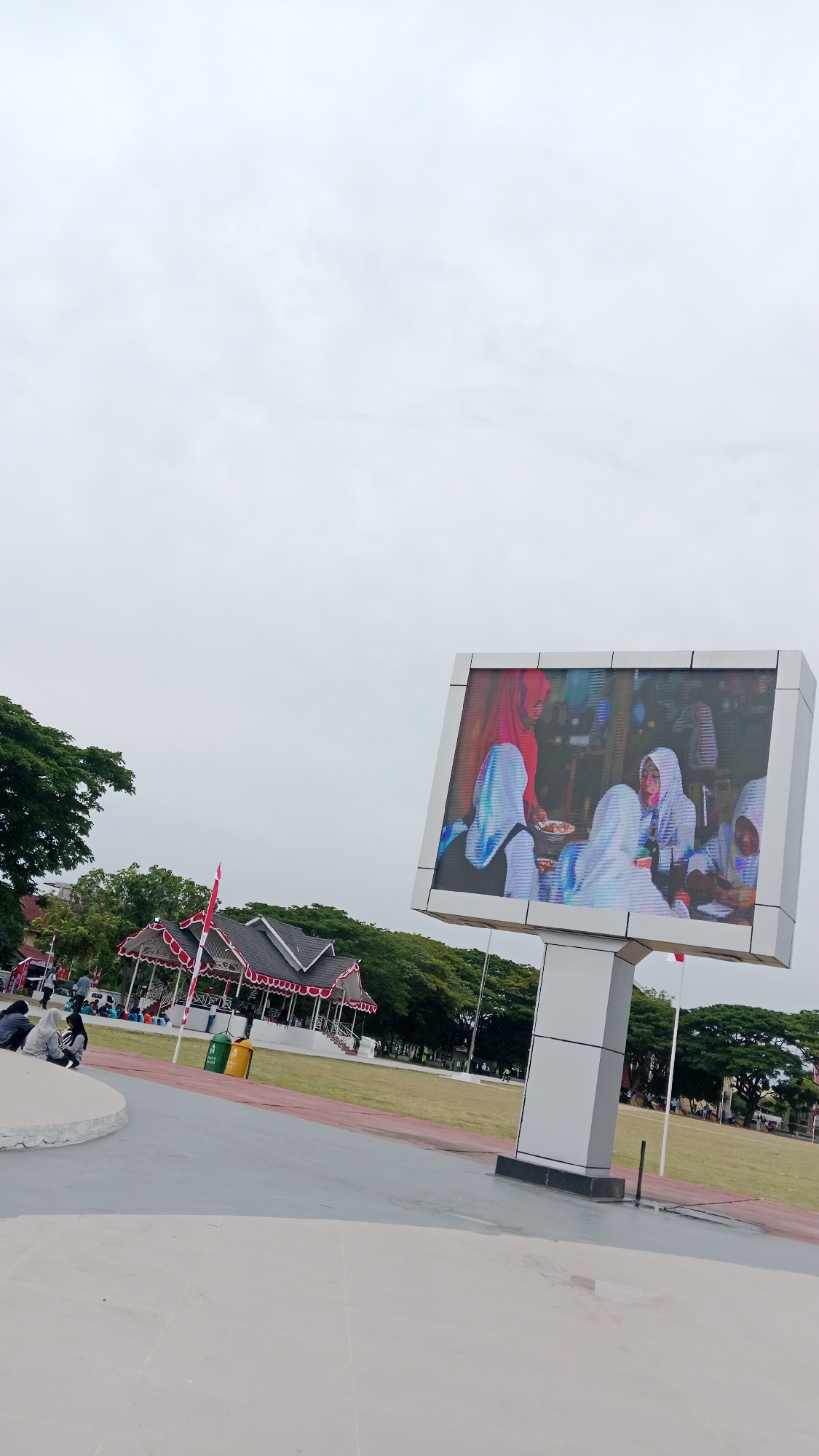 A large outdoor LED screen displays an image of several people wearing hijabs, with one person in a red hijab holding food. The screen is mounted on a white pillar and is located in a public outdoor space. In the background, there is a traditional Indonesian-style building with a dark roof and white decorative trim. Several trees are visible behind the building, and a grassy field stretches out in front of it. A few people are sitting on a circular concrete platform in the foreground, and others are scattered across the field. Two trash cans, one green and one yellow, are placed near the building. The sky is overcast. This scene is likely in Lhokseumawe, Indonesia, given the cultural elements visible.