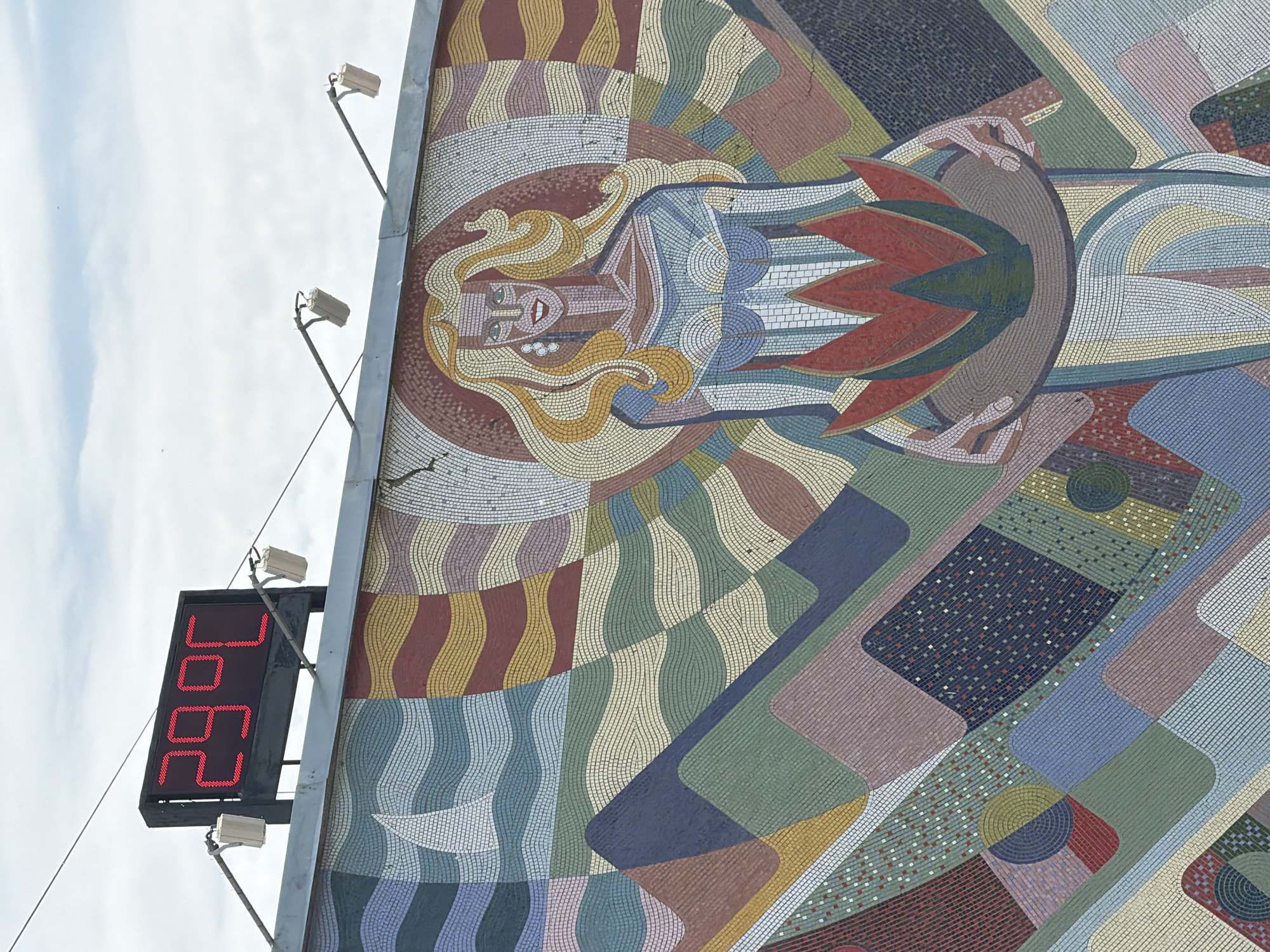 A medium shot captures a segment of a mosaic mural, seemingly affixed to a building wall in Kamyshin, Russia. A red LED display adjacent to the mural shows "26.9°C". The mosaic features a woman with blond hair, enclosed in a circular design. A sun-like element with red and green rays surrounds her. The mosaic's background consists of geometric shapes in various colors, including red, yellow, green, and blue. The sky is visible above the mural, indicating an outdoor setting. The image appears to be taken during daylight.