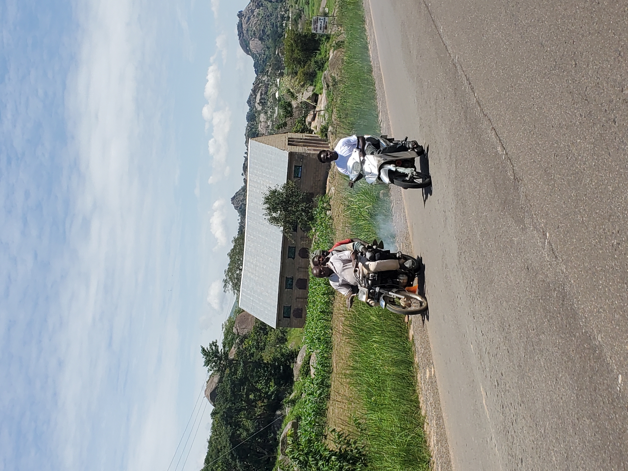 In Pankshin, Nigeria, two motorcycles are traveling on a paved road. On the left, a motorcycle carries three men; the driver is wearing a light-colored collared shirt and light pants, and is looking towards the camera. The two passengers behind him are partially obscured. On the right, a man wearing a white t-shirt and dark pants is driving a white scooter, also looking towards the camera. Both vehicles appear to be in motion, with smoke emanating from the exhaust of the motorcycle on the left. The road is bordered by a grassy verge and a field of crops. In the background, a building with a corrugated metal roof stands among trees and large rock formations, with a mountainous landscape visible beyond. The sky is partly cloudy.