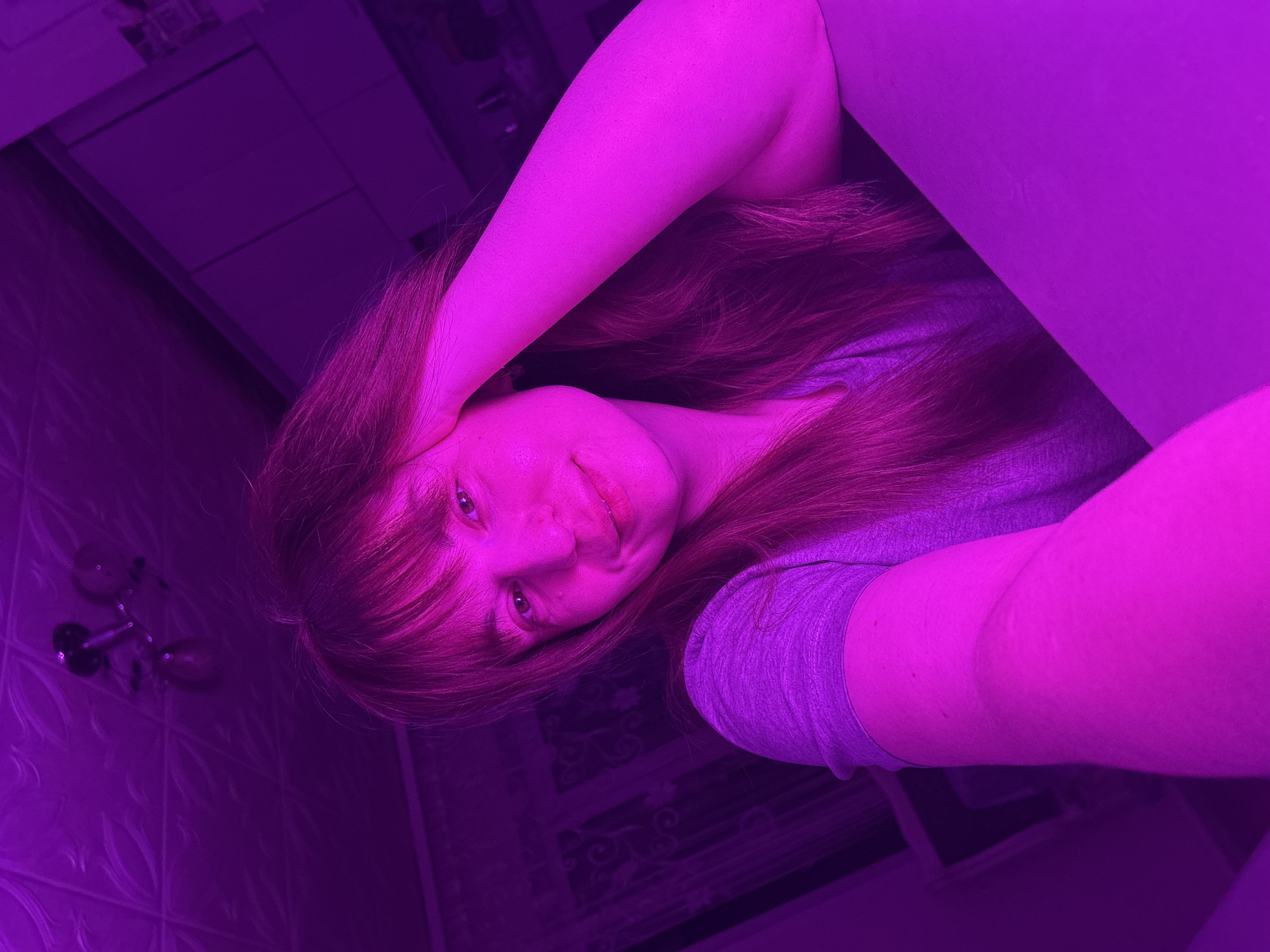 A casual selfie captures a young person with long, reddish-brown hair and bangs, looking directly at the camera with a subtle smile. They are wearing a light-colored (likely grey) t-shirt. One arm is raised, with the hand seemingly resting near the head, while the other arm is visible lower down.
The entire indoor scene is enveloped in a dominant purple and magenta light, which deeply hues all elements, making skin appear bright pink and hair a rich magenta. In the upper-left background, obscured by the light, are fixtures resembling a faucet and handles, along with a tiled wall, suggesting a bathroom or utility area. To the right, a piece of furniture, possibly a cabinet with drawers, is dimly visible, alongside a patterned surface that could be flooring or a rug.
The image is taken from a slightly low angle, with the subject positioned diagonally, indicative of a handheld shot. The person's expression is relaxed and friendly. No specific time of day can be determined due to the pervasive artificial lighting. There is no visible text in the image. The location context provided is Фоки, Russia.