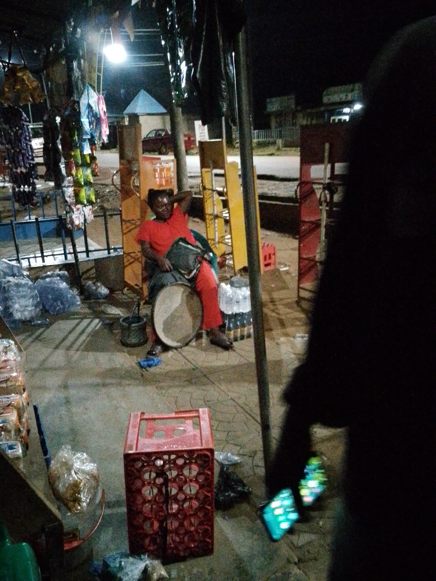 A person wearing a red outfit sits with their hand behind their head, resting on a dark-colored drum. A bag is slung over their shoulder. The person is sitting on the ground next to a large, round, flat object, possibly a platter or tray, with a rim. Behind the person, various items are displayed, including what appear to be bags of snacks hanging from a structure. Several large, clear plastic bags filled with items are on the ground to the left of the person. A red plastic crate with an intricate pattern is visible in the lower right of the image, and a black bucket is near the person. The scene appears to be outdoors at night, with artificial lighting illuminating parts of the area. In the background, a red car is visible. The ground is paved.