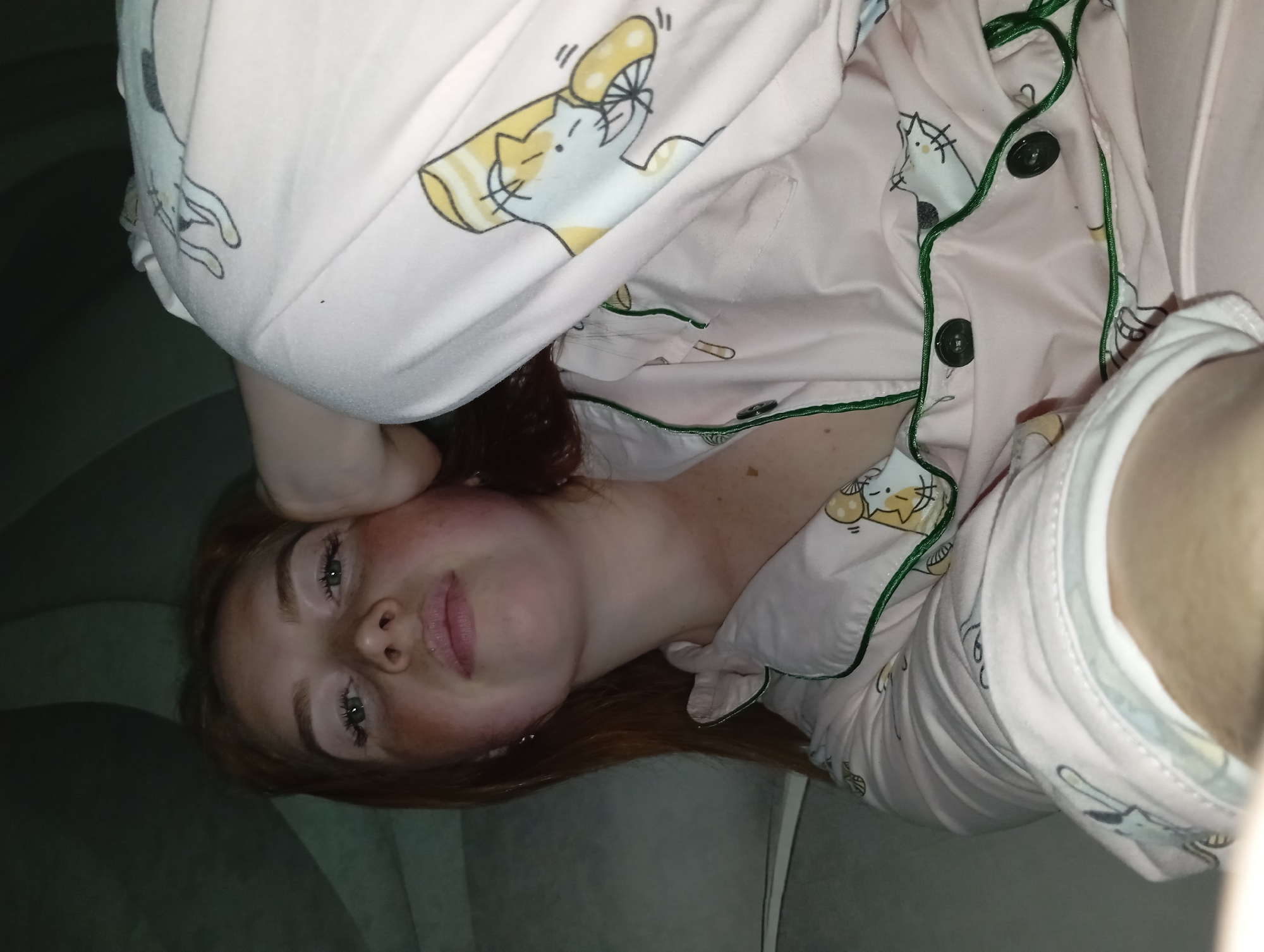 A red-haired woman wearing pink pajamas with a cartoon cat print lies on a gray sofa. The pajamas have green piping and dark buttons. The woman's face is visible, with gray eyes and fair skin, slightly flushed, and the photo appears to be taken in low lighting conditions. The location in Yurgamysh, Russia is not discernible from the image.