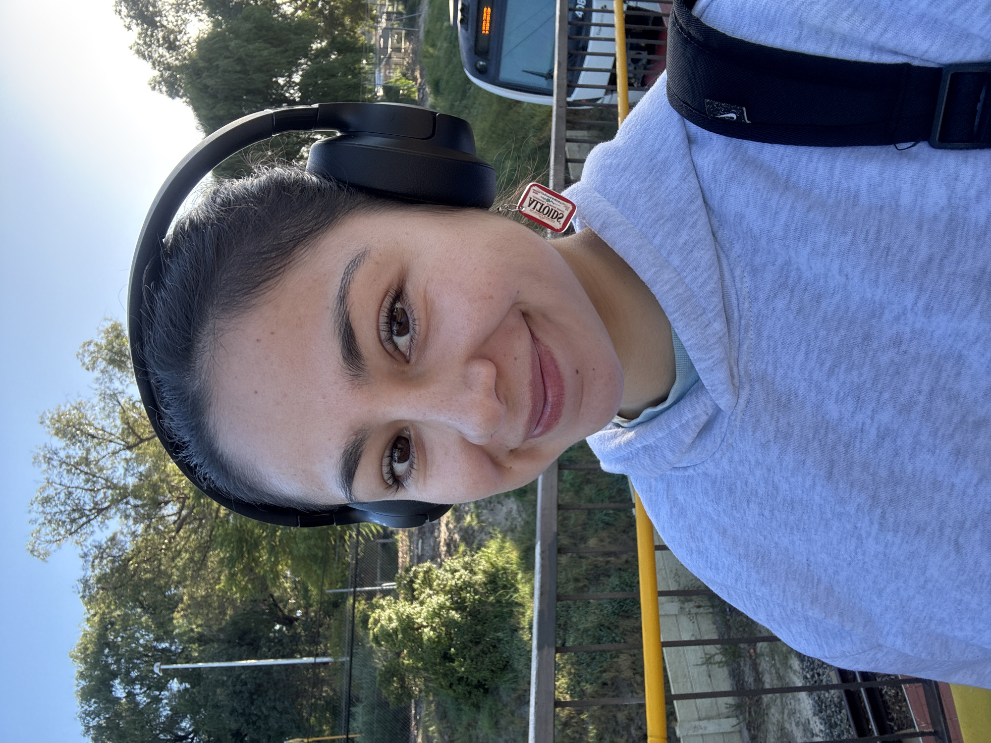 A person stands on a platform, likely at a train station in Bassendean, Australia. They are wearing black headphones, a light grey hooded sweatshirt, and a black backpack with a visible Nike logo. A red tag with reversed writing hangs off the headphones, near the person's left ear. The person is looking towards the camera with a slight smile. The environment includes a train in the background, trees, a fence, and yellow railing. The location is consistent with a train station in Bassendean, Australia, considering the type of train and surrounding vegetation.