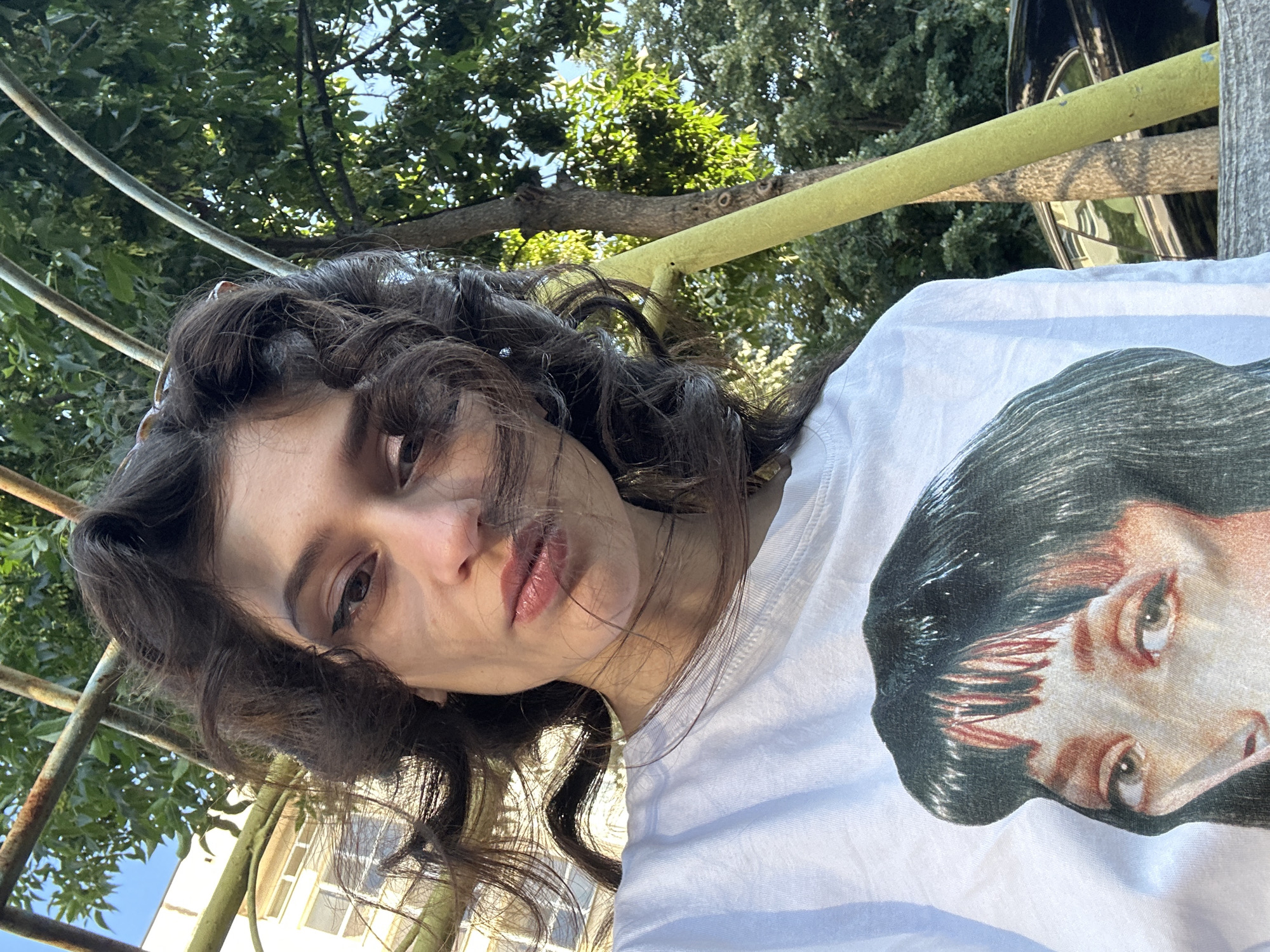A young woman with dark, curly hair and dramatic eye makeup poses for a close-up selfie outdoors. She is wearing a white t-shirt with a graphic print of a woman's face. Her hair is windswept, partially obscuring her face. Behind her, a building with white walls and windows is visible, along with lush green trees and a yellow pole. The lighting suggests it is daytime. The scene appears to be in an urban or semi-urban setting, possibly a park or courtyard. The location is identified as Sofia, Bulgaria, on "G. M. Dimitrov" Boulevard.