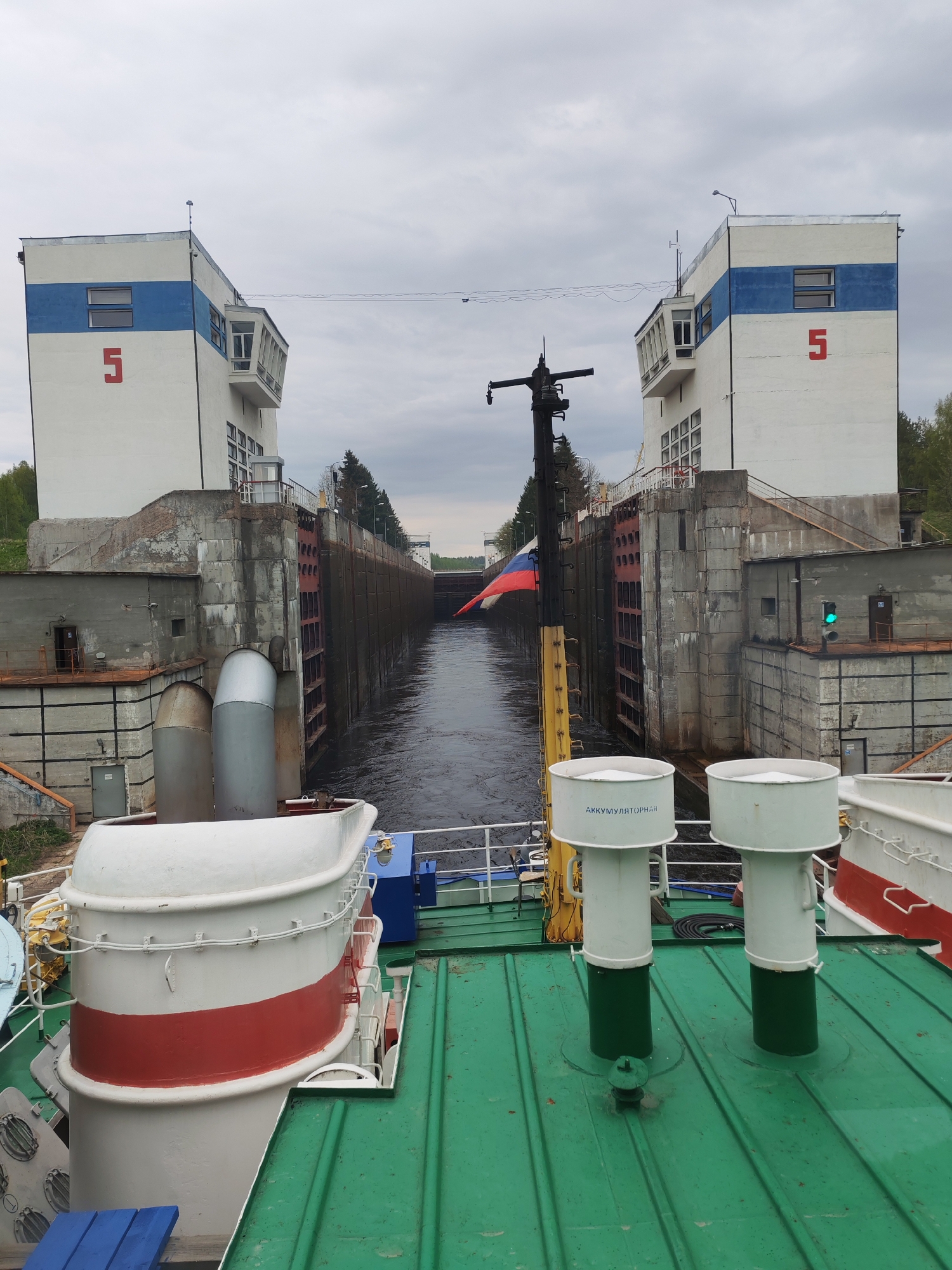 A view from the deck of a ship passing through lock 5. The lock's towers are white with blue stripes and a large red number "5" on each. The Russian flag is flying from a mast on the ship. The water is dark and the sky is overcast.
