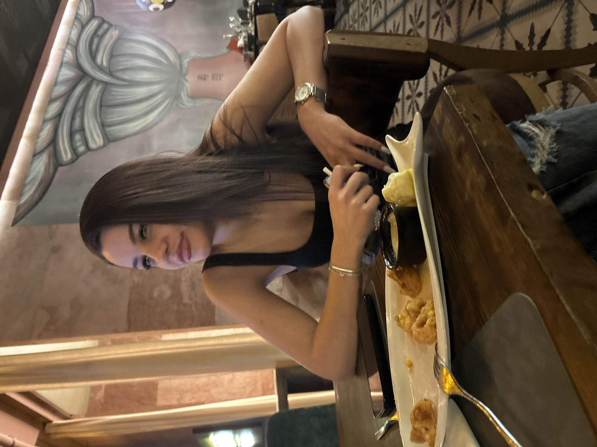 A young woman sits at a table in a restaurant in Plovdiv, Bulgaria. She wears a black tank top and a silver wristwatch. Her dark hair is styled straight. She holds a small utensil, possibly a toothpick or skewer, over a white serving dish containing fried food and a dipping sauce. On the table, there are also eating utensils. The background reveals the restaurant's interior, featuring a mural on the wall and a patterned tile floor. The overall lighting suggests an indoor setting.