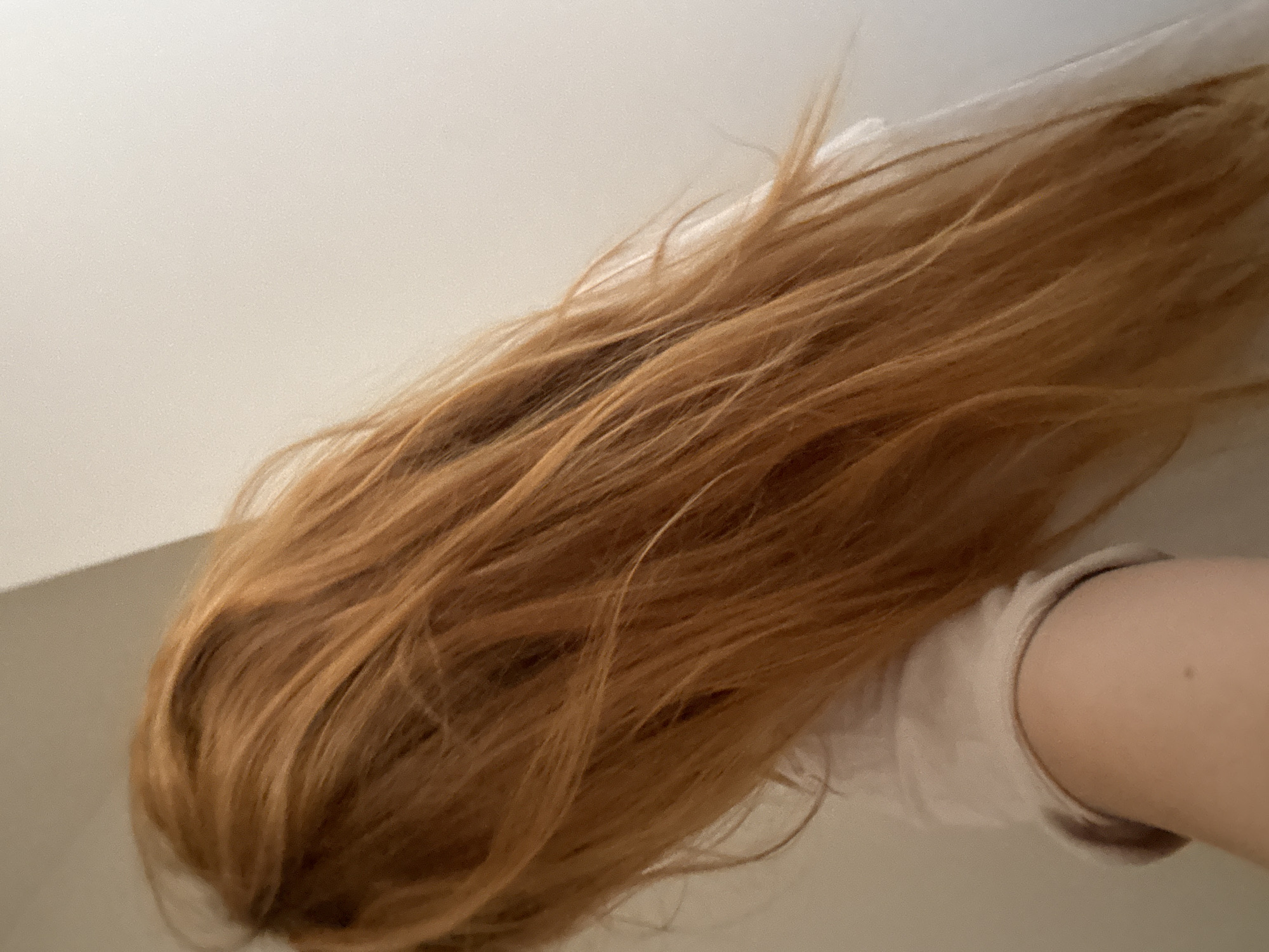 The image shows a close-up of a person's long, auburn hair against a plain, light-colored background. The hair is loose and appears to be slightly windswept. A small portion of the person's shoulder and upper arm, wearing a light-colored garment, is visible on the left side of the frame. The scene is indoors, with no discernible features of Budva, Montenegro.