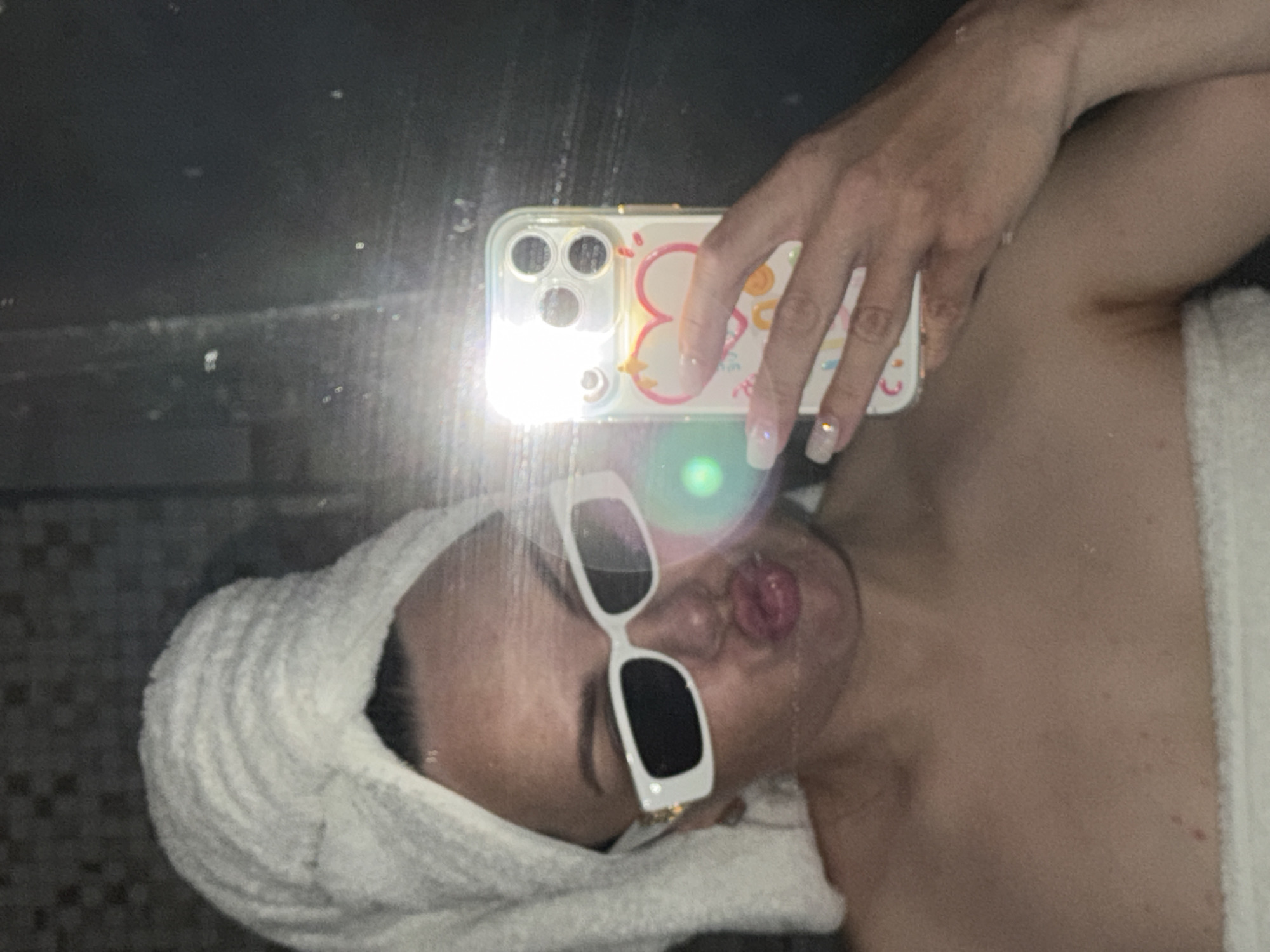 A person with a towel wrapped around their head is taking a selfie in what appears to be a bathroom setting. The person is wearing white-framed sunglasses and has their mouth pursed. They are holding a smartphone with a decorative case. The background shows a tile wall, likely within a bathroom. The location in the city of Votkinsk, Russia, cannot be confirmed from the image alone.