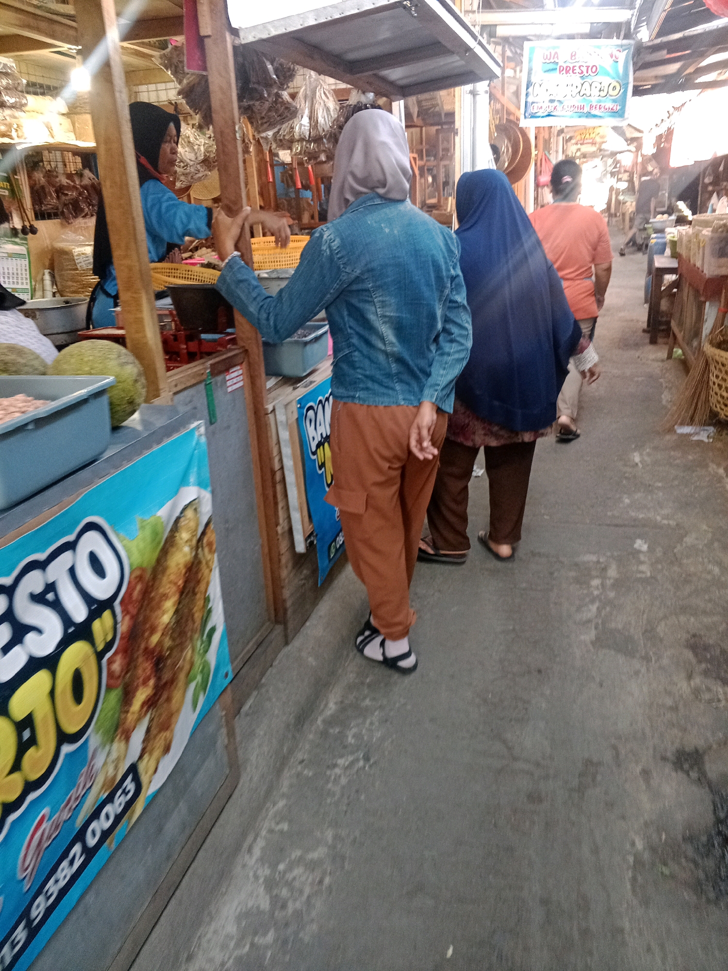 The image depicts a street scene, likely in Wonogiri, Indonesia, with a market atmosphere. In the foreground, a person wearing a denim jacket and brown cargo pants is walking away from the camera. They have on black and white sandals. To their left, a blue banner with the word "PRESTO" and an image of fried fish is prominently displayed. Further to the left, a woman wearing a blue hijab and tunic is visible behind a stall, interacting with someone. In the background, several other individuals are seen walking along the street, with stalls and buildings lining the path. The ground appears to be paved with concrete. There is a sign above the stalls that reads "WA B. PRESTO KEMBARJO". The overall lighting suggests it is daytime.