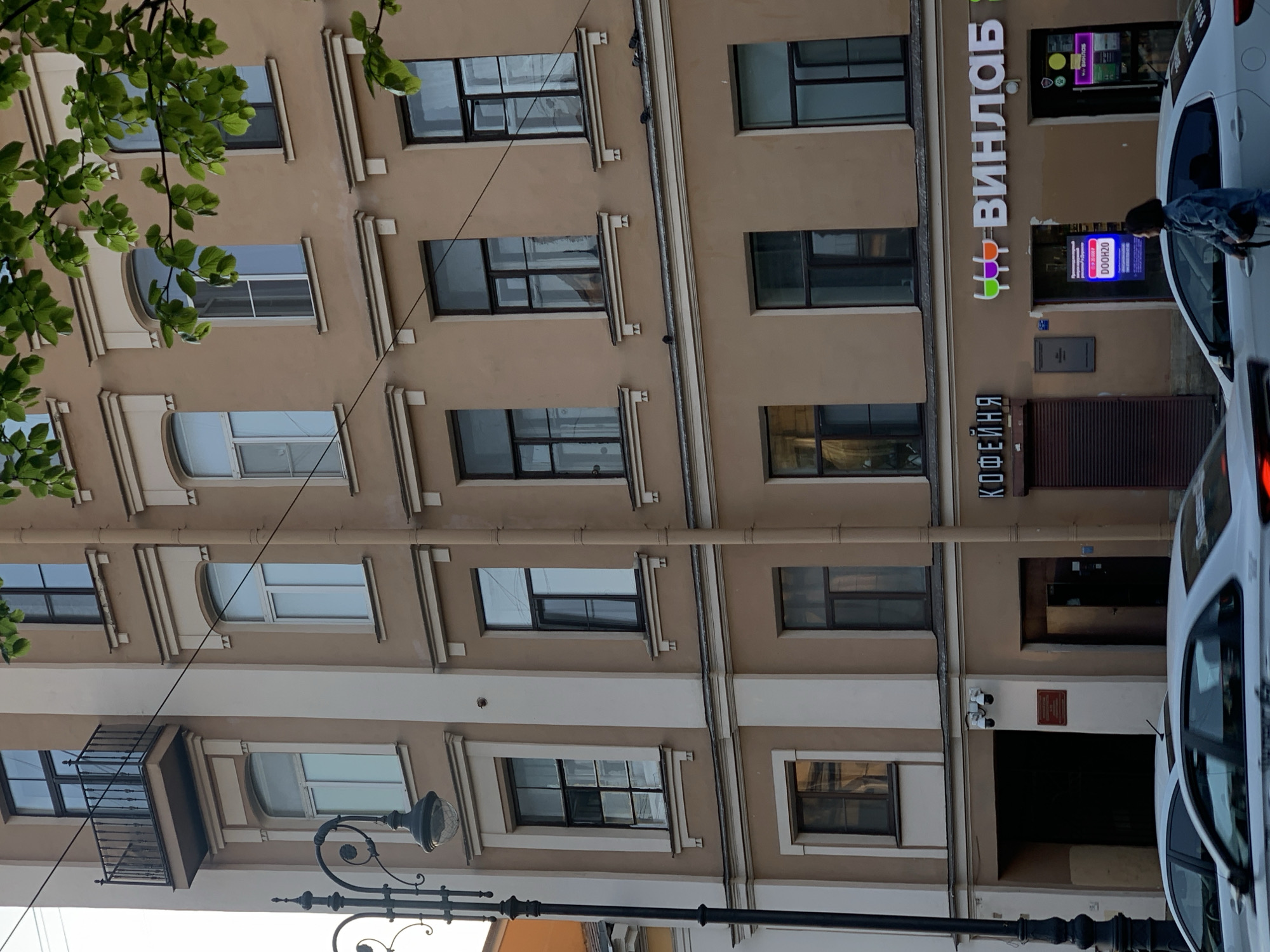 The photograph depicts a building in Saint Petersburg, Russia. The building has a tan facade with white trim around the windows and decorative vertical elements. The windows are rectangular and uniformly spaced. The lower left part of the image features green leaves from a tree. A section of an ornate black streetlight is present in the lower part of the frame. On the right side of the building, there is a sign that reads "ВИНЛАБ" (Vinlab) vertically and "КОФЕЙНЯ" (Coffee Shop) below it. The entrance is closed. A partial view of a white vehicle is obstructing the view of the entrance. A person is near the white vehicle.