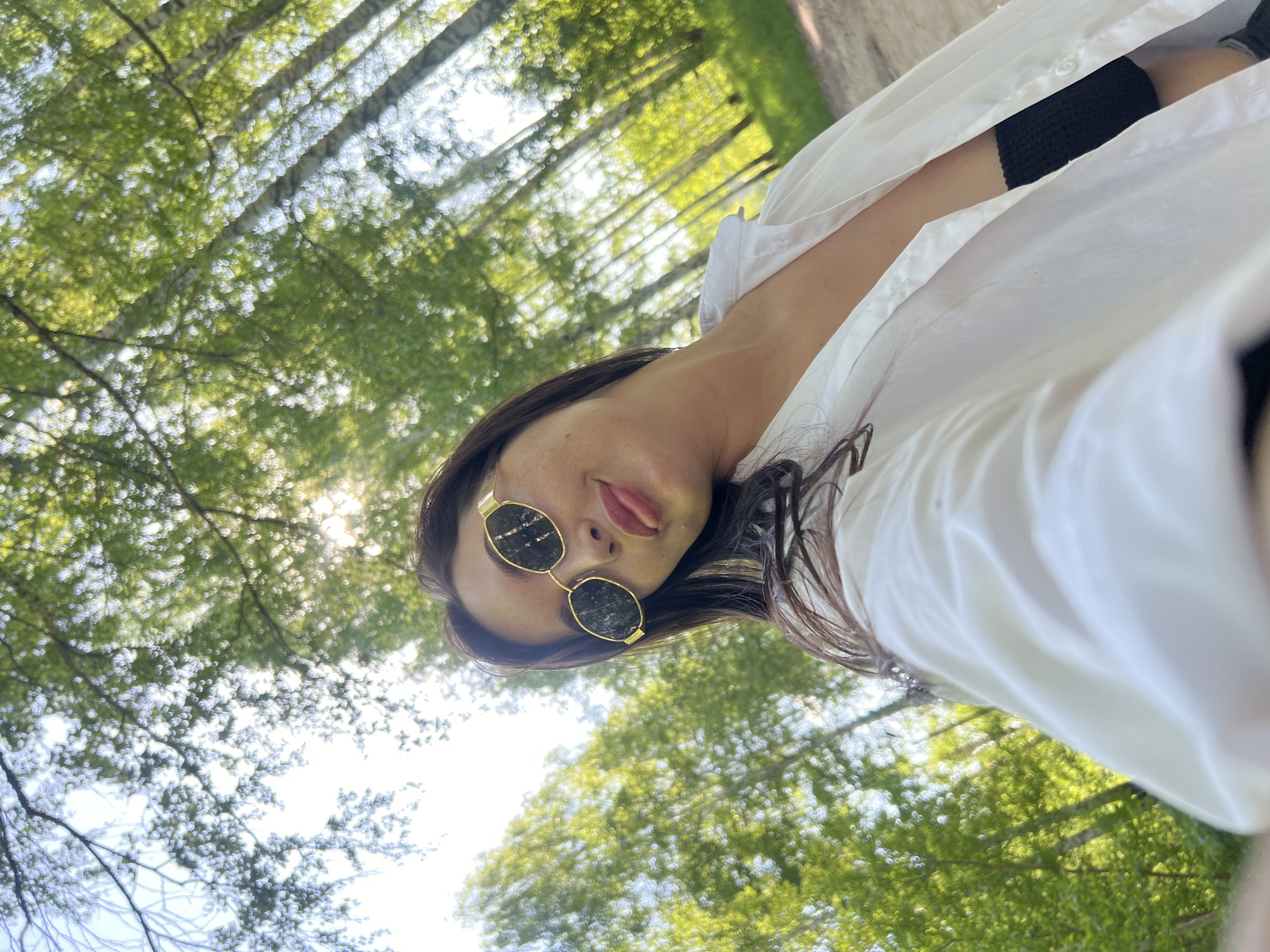 A woman is seen in a selfie with green trees and a blue sky in the background. She wears sunglasses with gold frames and dark lenses, and a white button-down shirt with a black garment visible underneath. She appears to be outdoors in a wooded area, but the exact location within Chaikovsky, Russia is not discernible.