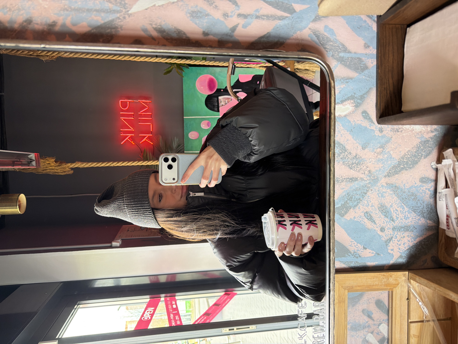 A young woman is captured in a mirror selfie, seen from the chest up, inside what appears to be a cafe in Khimki, Russia. She has long, dark hair and is wearing a grey knit beanie and a black puffer jacket. Her expression is neutral, and she is looking directly at the camera of the light-colored smartphone she holds up to take the photo. In her other hand, she holds a white take-away cup patterned with the word "PINK" in pink letters.
Reflected in the mirror behind her is a dark wall featuring a red neon sign that spells "PINK" (appearing backward due to the reflection). To the right of the sign, a framed artwork on a green background depicts a stylized figure with pink circular elements. A thick rope is visible horizontally strung across the wall. To the left, a golden cylindrical object stands on a surface.
Outside the mirror, the wall on the right is decorated with an abstract pattern in light pink and blue hues. Wooden shelving units are built into this wall, one holding white napkins and another containing small white packets (likely sugar or creamers). The lower left corner of the image shows a glass door or window with red tape across it. Visible text on the tape includes "OPEN" and "MILK CAFE," along with other small, partially obscured details. The overall setting suggests a cozy, modern cafe environment.