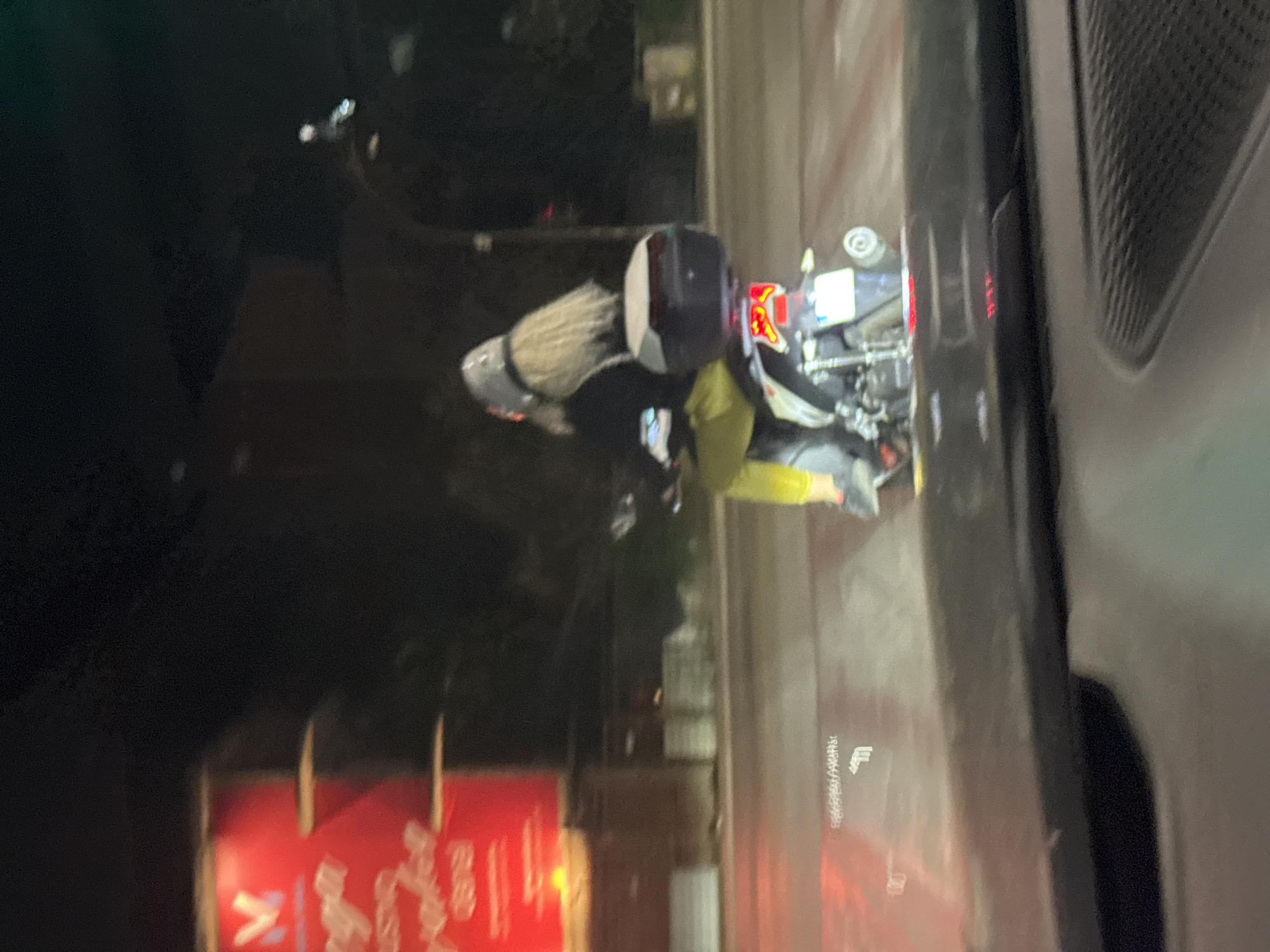 A person with long blond hair, wearing a black shirt, yellow pants, and a silver helmet, rides a scooter at night. The scooter has a black storage box on the back. The scene appears to be on a street in Budva, Montenegro, with a red illuminated sign in the background. The camera is positioned inside a vehicle, capturing the scooter from behind and slightly to the side.