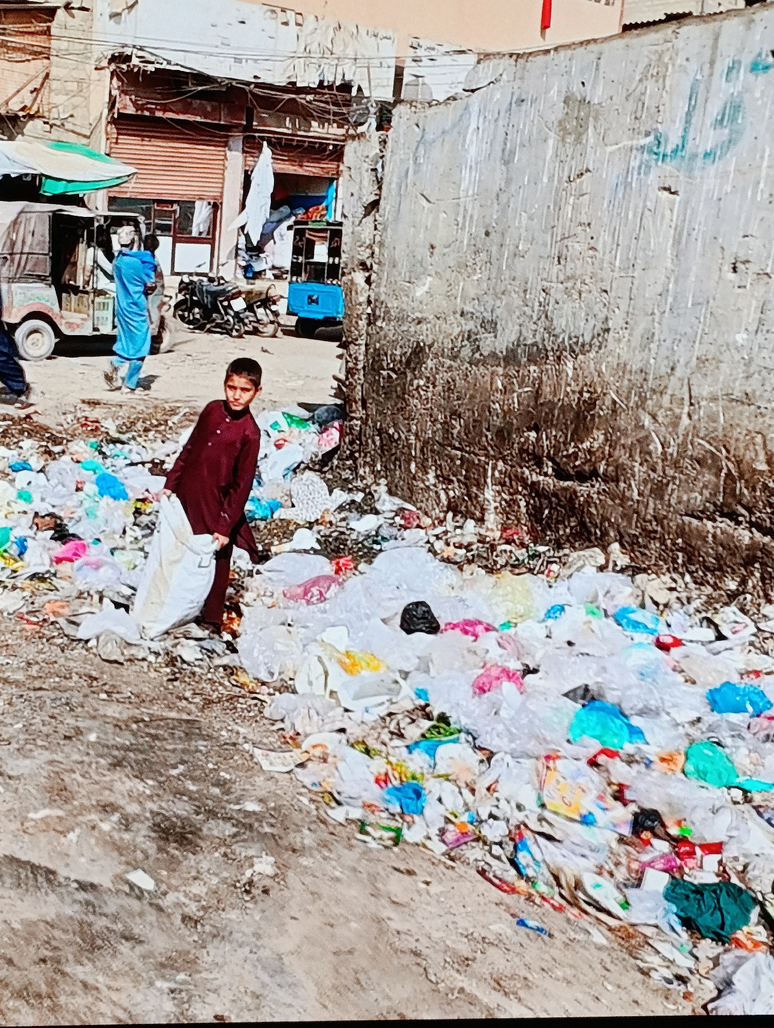 In the city of Unknown, Unknown, the image depicts a boy standing among a pile of garbage on a street. He is wearing a maroon outfit and holding a white sack. Behind him, there is a concrete wall and in the background, there are buildings, a motorcycle, and motorized vehicles. Another person, wearing a blue robe, is walking in the background as well. The street is dirty and littered with trash.