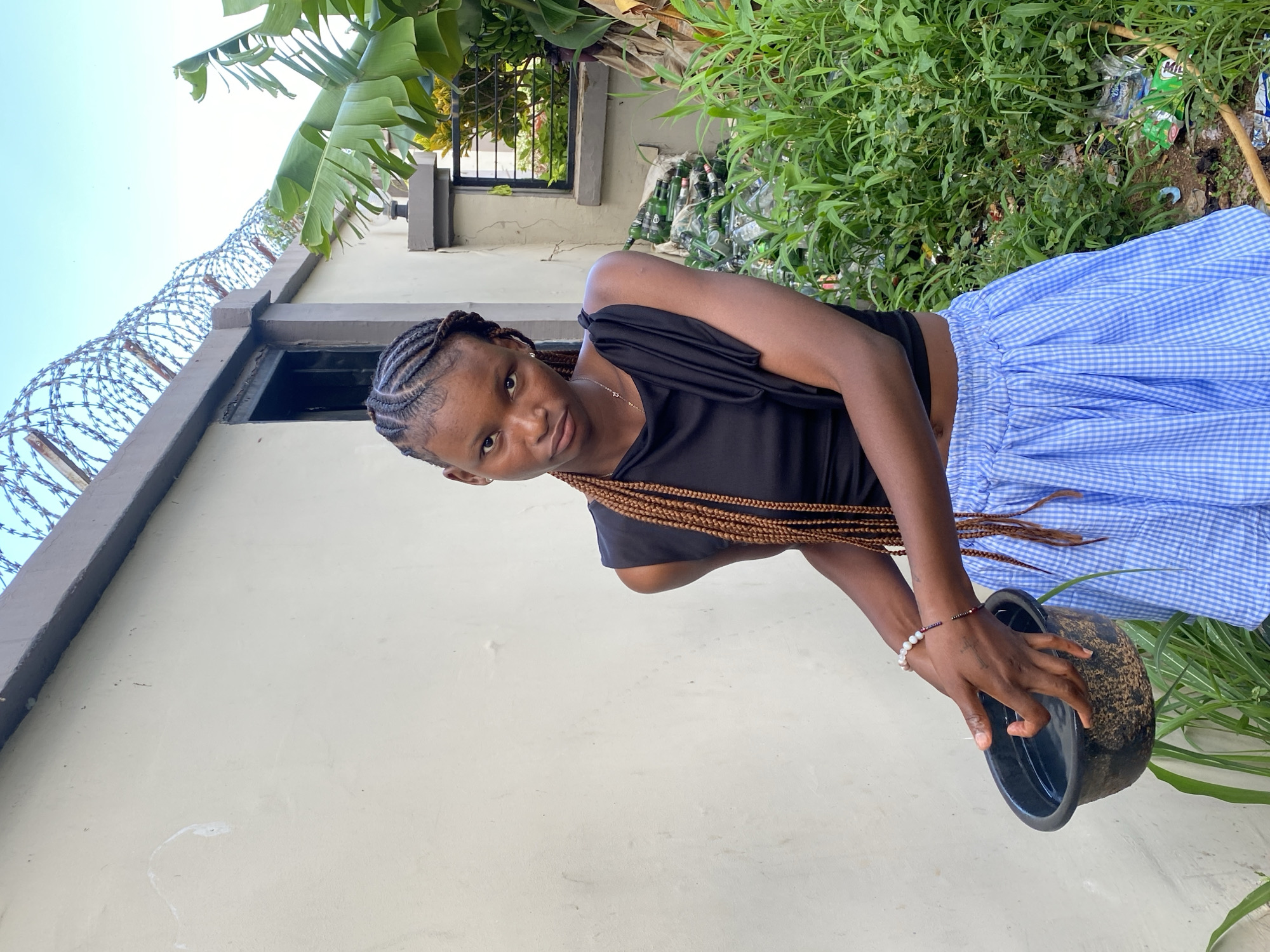 In Gura Top, Nigeria, a young Black woman with braided hair stands against a light-colored wall. She is wearing a black, off-the-shoulder top and a blue and white checkered skirt. She is holding a black bowl in her hands, which appear to be submerged in the water within the bowl. The background shows a wall topped with barbed wire, lush green foliage, and a pile of green glass bottles. The sky above is a clear blue.