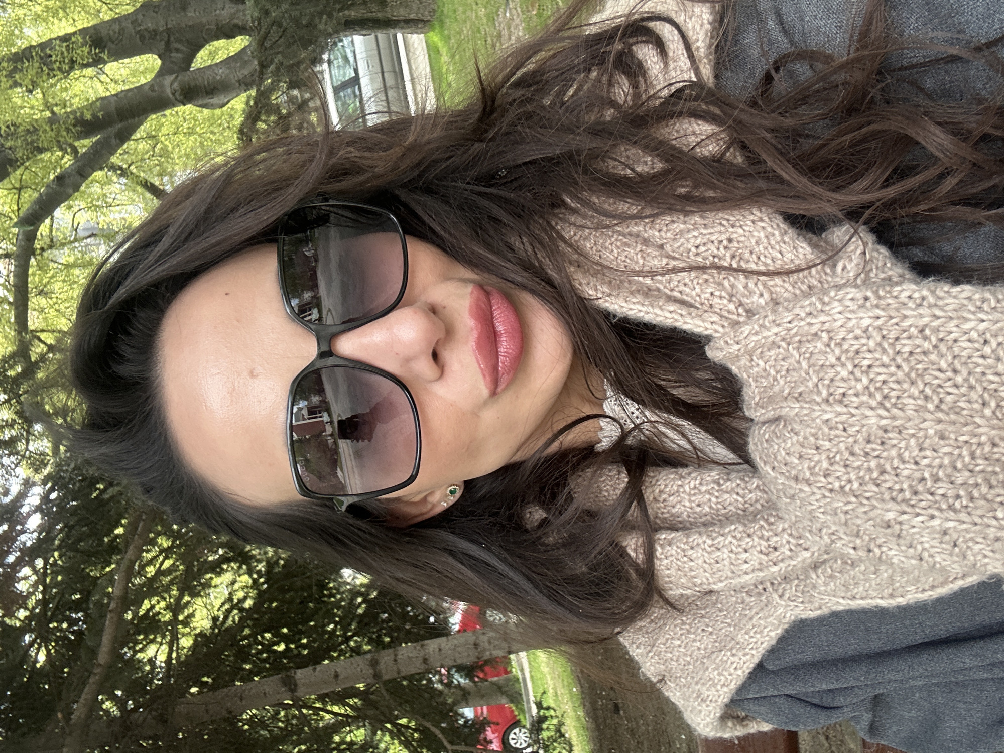 A woman is seen in a portrait shot, wearing sunglasses, a gray garment, and a beige knitted scarf. The background contains trees and greenery, with a glimpse of a red car and a building. The scene appears to be outdoors, possibly in a park setting. There's no specific indication of Varna, Bulgaria.