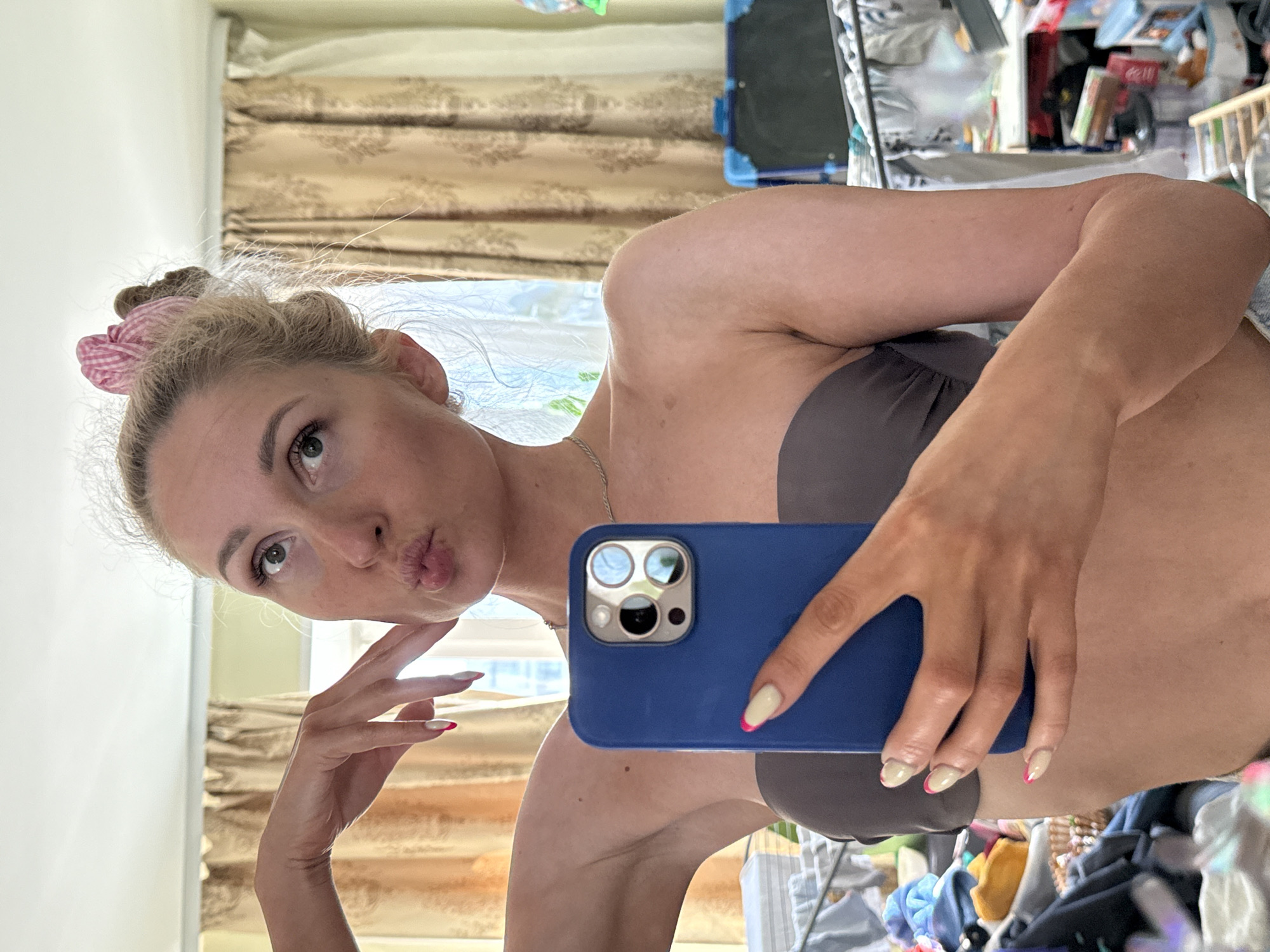 A woman is taking a selfie indoors, holding a blue iPhone 13 Pro Max. She has fair skin, blonde hair tied up with a pink and white checkered scrunchie, and is wearing a gray strapless bra. Her fingernails are painted with a nude color and a pink accent at the tips. She is pursing her lips for the selfie.
Behind her are beige patterned curtains and a window. The background also shows a cluttered space with various objects, including a suitcase, books, and clothes. The location is unspecified and the city of Chaikovsky, Russia is not identifiable in the image.