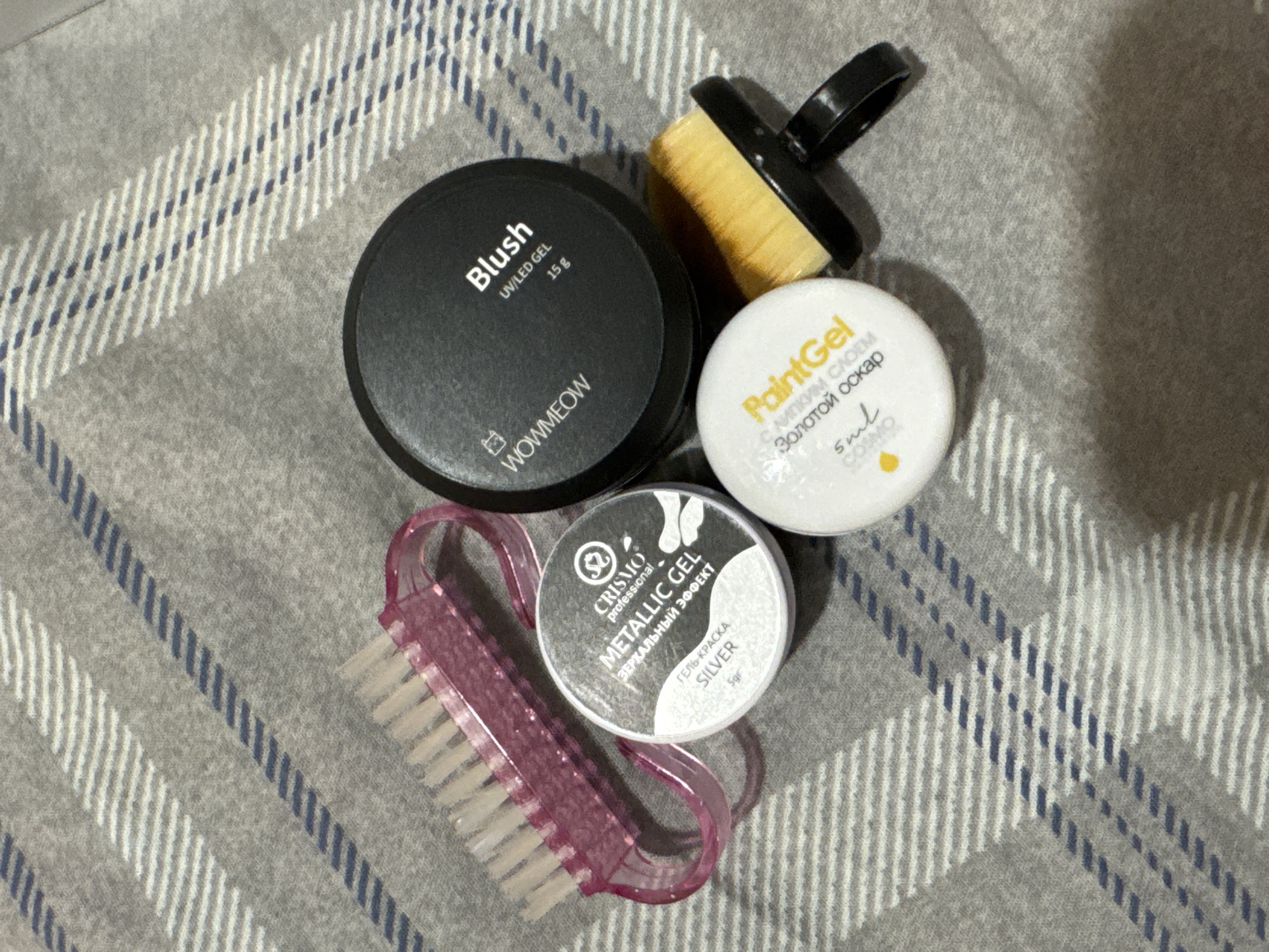 The image displays a close-up, overhead view of several nail art products arranged on a grey, checkered fabric. The objects include two nail brushes, one pink and translucent with a curved handle, and another with a black handle and yellow bristles. There are also three small containers of nail gel. One is a black container labeled "WOWMEOW Blush UV/LED GEL 15g". Another is a white container with a black lid, labeled "CRISMO PROFESSIONAL METALLIC GEL ЗЕРКАЛЬНЫЙ ЭФФЕКТ SILVER 5gr". The third is a white container with yellow lettering that reads "PaintGel Золотой оскар 5ml". The background fabric is a soft grey material with a pattern of alternating thick white and thin blue stripes running diagonally across it. The lighting is even, suggesting an indoor setting. No people are visible in the image. The location in the city of Яромінскі сельскі Савет, Belarus is not discernible from the visual information provided.