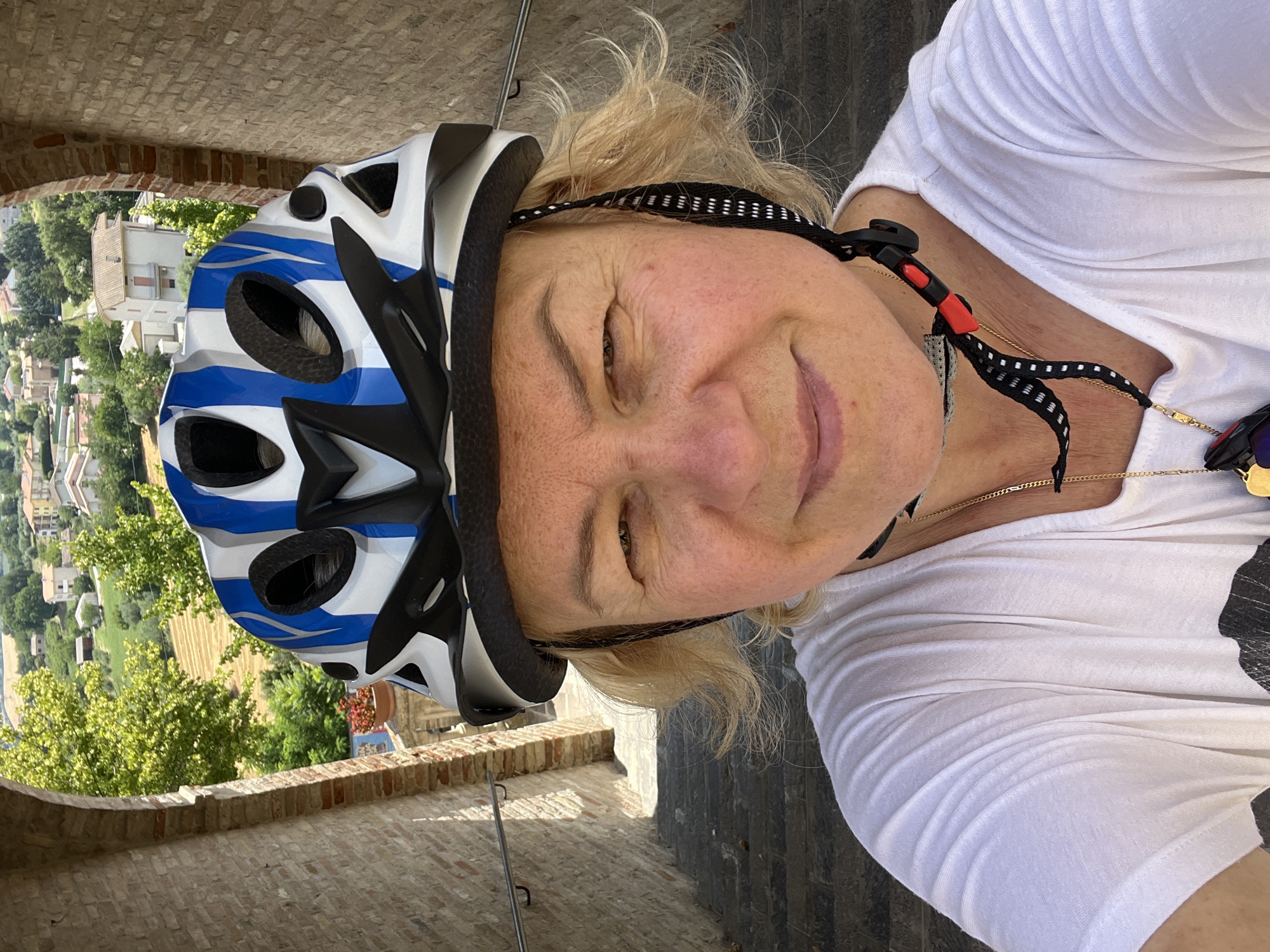 A woman wearing a white t-shirt and a blue and white bicycle helmet is taking a selfie on a stone staircase. The woman has blonde hair and is smiling. The background shows a view of houses and trees in the city of Barbara, Italy. The staircase is made of stone and has a metal railing on the left side. The woman is wearing a gold necklace.