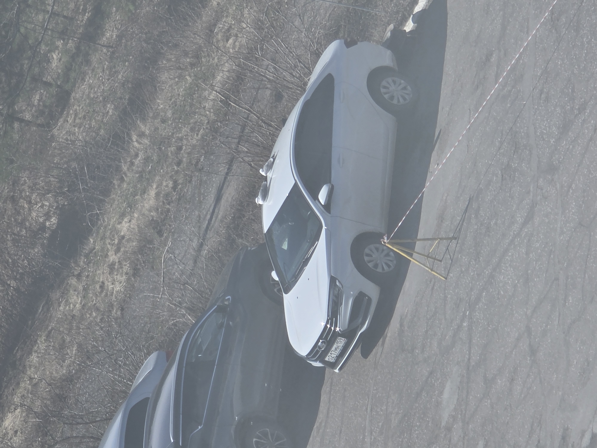This image, captured from an elevated perspective and rotated, depicts an outdoor scene in what appears to be a parking area or street in Chaikovsky, Russia.
The primary subject is a white sedan, prominently parked on an asphalt surface. This car is identifiable as a Lada Vesta, distinguished by its front grille and badging. A Russian license plate, "O217MM 159", is clearly visible on its front bumper. The "159" region code corresponds to Perm Krai, confirming the location context. Partially visible behind and to the left of the white car is a second vehicle, a dark-colored sedan or hatchback.
Adjacent to the front left wheel of the white car, a yellow metal structure resembling a foldable barrier or stand is placed on the asphalt. Extending from this structure is a red and white striped warning tape, which stretches across the asphalt surface, suggesting an area of demarcation, temporary restriction, or caution.
To the left of the parked cars, the ground transitions into a natural, unkempt area. This section features numerous bare tree branches and dry, light-colored grass or brush, indicating either late winter or early spring. The overall lighting suggests daytime, possibly under hazy or overcast skies, creating soft shadows and a slightly muted color palette. No people are visible in the frame, and no specific activity beyond the parking of vehicles and the presence of the barrier is evident.