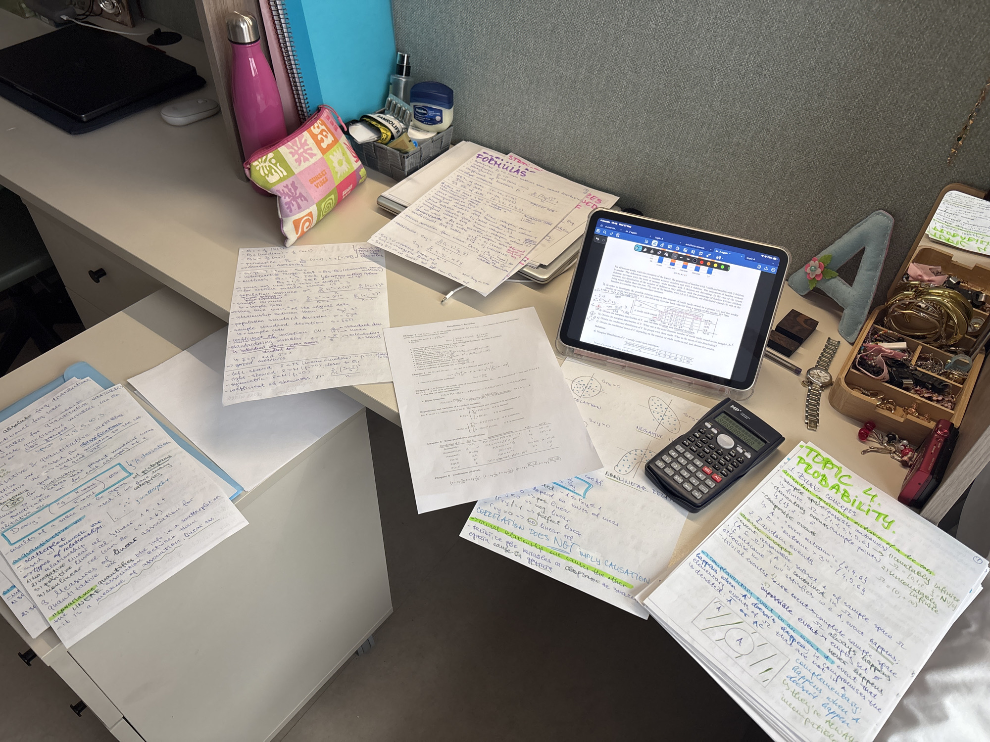 The image shows a desk cluttered with study materials. There are handwritten notes on various topics, some in blue folders. A tablet displays a document, likely related to the notes. A calculator sits near the tablet. Other items on the desk include a water bottle, a pouch, and a jewelry box. The environment suggests a study area, possibly in a home. No specific location within Getafe, Spain is evident from the image.