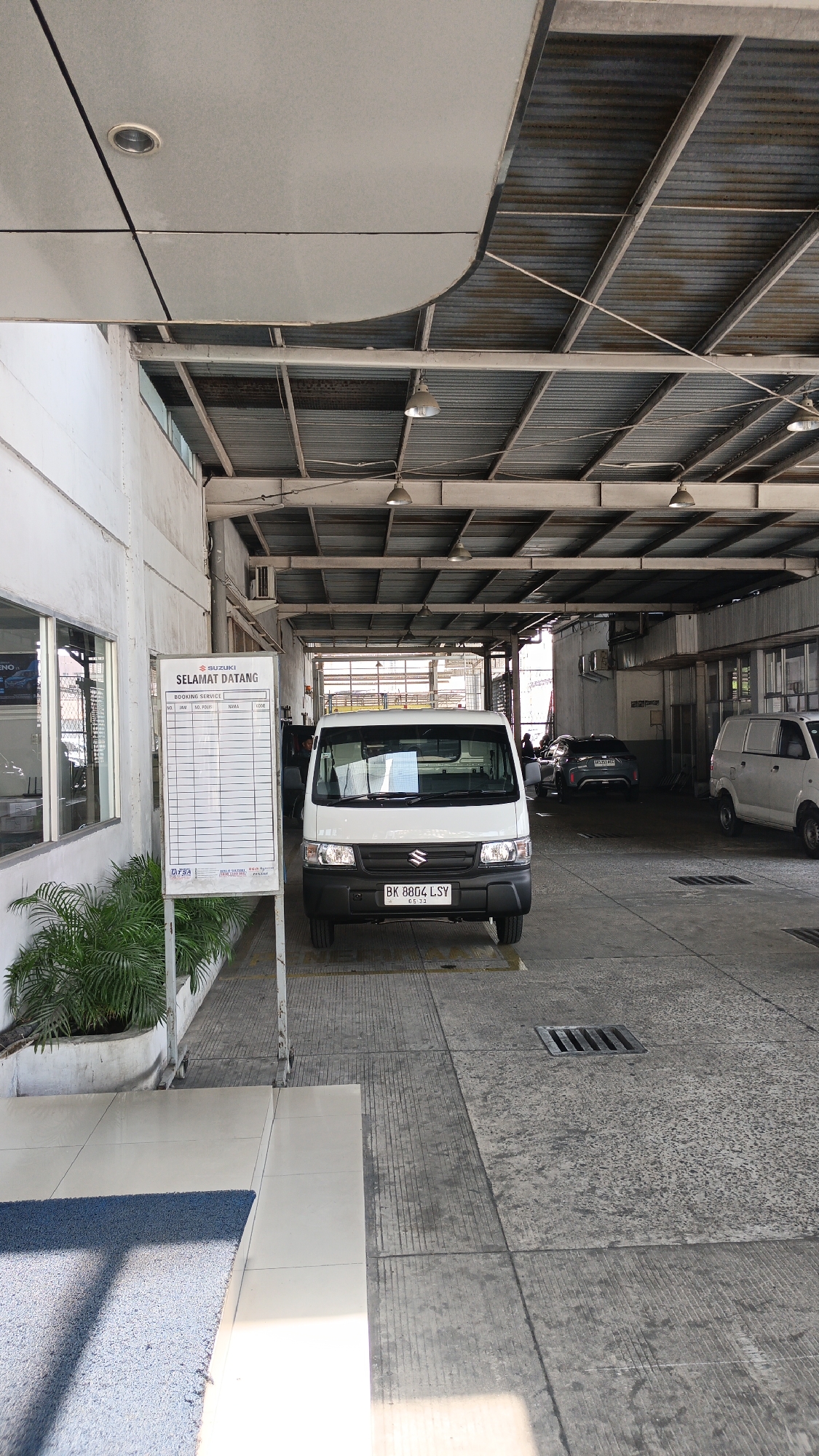 An outdoor view of what appears to be a car repair or service bay in Medan, Indonesia. In the foreground, a white Suzuki Carry van with license plate "BK 8804 LSY" is parked in front of a white reception desk/signage. The signage contains text in Indonesian, including "SELAMAT DATANG" (Welcome) and "BOOKING SERVICE." The environment is utilitarian, with a corrugated metal roof supported by metal beams and exposed lighting fixtures. Concrete flooring with visible expansion joints and a drainage grate is present. Lush green plants are in planters to the left of the van. In the background, other vehicles, including a dark-colored sedan and a white van, are visible within the service bay area. The scene suggests a commercial establishment focused on vehicle services.