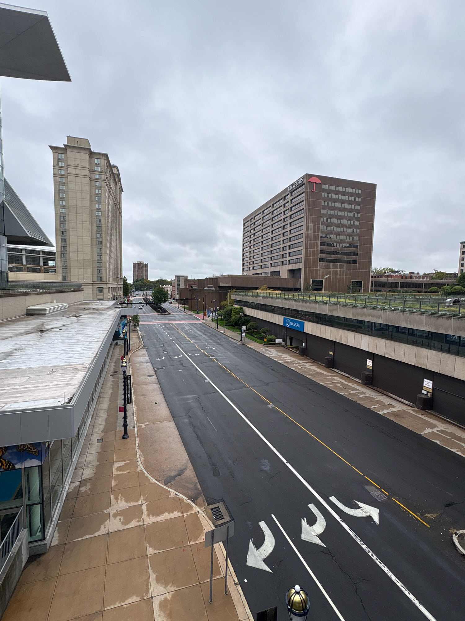 This is a high-angle, full-shot, outdoor photograph taken on an overcast day in Hartford, Connecticut, United States of America. The image captures a city street scene with several buildings and infrastructure elements.
In the foreground, a section of a light-colored building with a slanted roof occupies the top left portion of the frame. To the right of this, a wide asphalt road stretches into the distance, marked with white and yellow lines, and directional arrows indicating lanes for left turns and straight ahead travel. The road appears wet, suggesting recent rain. The sidewalks are paved with light brown square tiles, also showing signs of wetness. Various street lights and traffic control devices are visible along the sidewalk.
Dominating the middle ground and background are several multi-story buildings. On the left, a tall, light-colored building with numerous windows stands prominently. To its right, a larger, darker building with a visible "TRAVELERS" sign and a red umbrella logo at its top is a key feature. Below this building, a concrete structure with a green, landscaped edge runs horizontally, appearing to be an elevated walkway or part of a parking garage. A blue sign with the word "NASSAU" is affixed to this structure. Further into the background, more buildings of varying heights and architectural styles are visible, with trees and vegetation interspersed among them. The sky is completely obscured by thick, gray clouds. No people are clearly visible in the image. The overall impression is one of an urban environment during a period of subdued natural light.