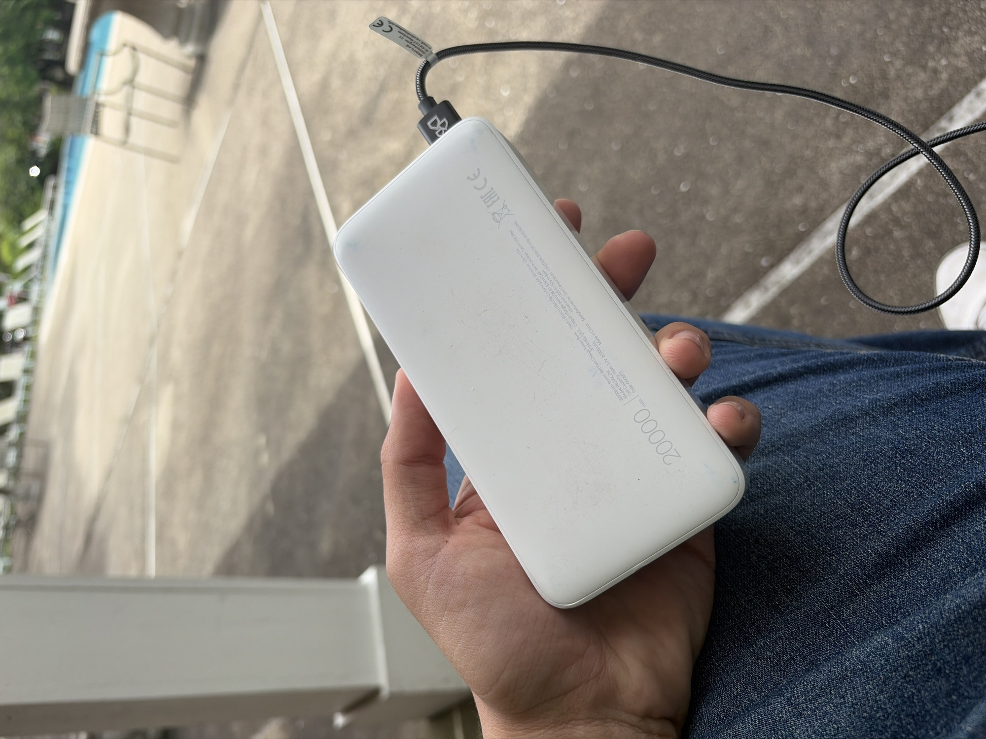 A white power bank, labeled "20000 mAh," is held in a hand over a pair of blue jeans. A black charging cable is plugged into the device. The background features a gray concrete area with architectural elements like a white railing. It is impossible to determine if the image was taken in Edison, United States of America.