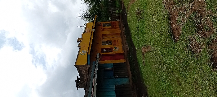 The scene is set in Gatakaine, Kenya, under an overcast sky. Two single-story buildings stand side-by-side. The structure on the left features a weathered blue wooden facade and a dark roof with a visible chimney or vent pipe. The adjacent, more prominent building on the right has a yellow-painted facade with a stepped parapet roofline. This yellow building contains four openings: a red door on the far left, two barred windows separated by a central red door, and another red door on the far right. Two distinct "CooP O" logos are visible on the yellow facade, one below the left window and another above the right section of the building. Faded text is present above the central door. A thin, dark utility line extends horizontally from above the right side of the yellow building. The foreground consists of green grass interspersed with patches of dark, exposed soil. To the right of the buildings, dense green trees and foliage are visible. No individuals, vehicles, or animals are present in the image, and no active interactions or actions are depicted.