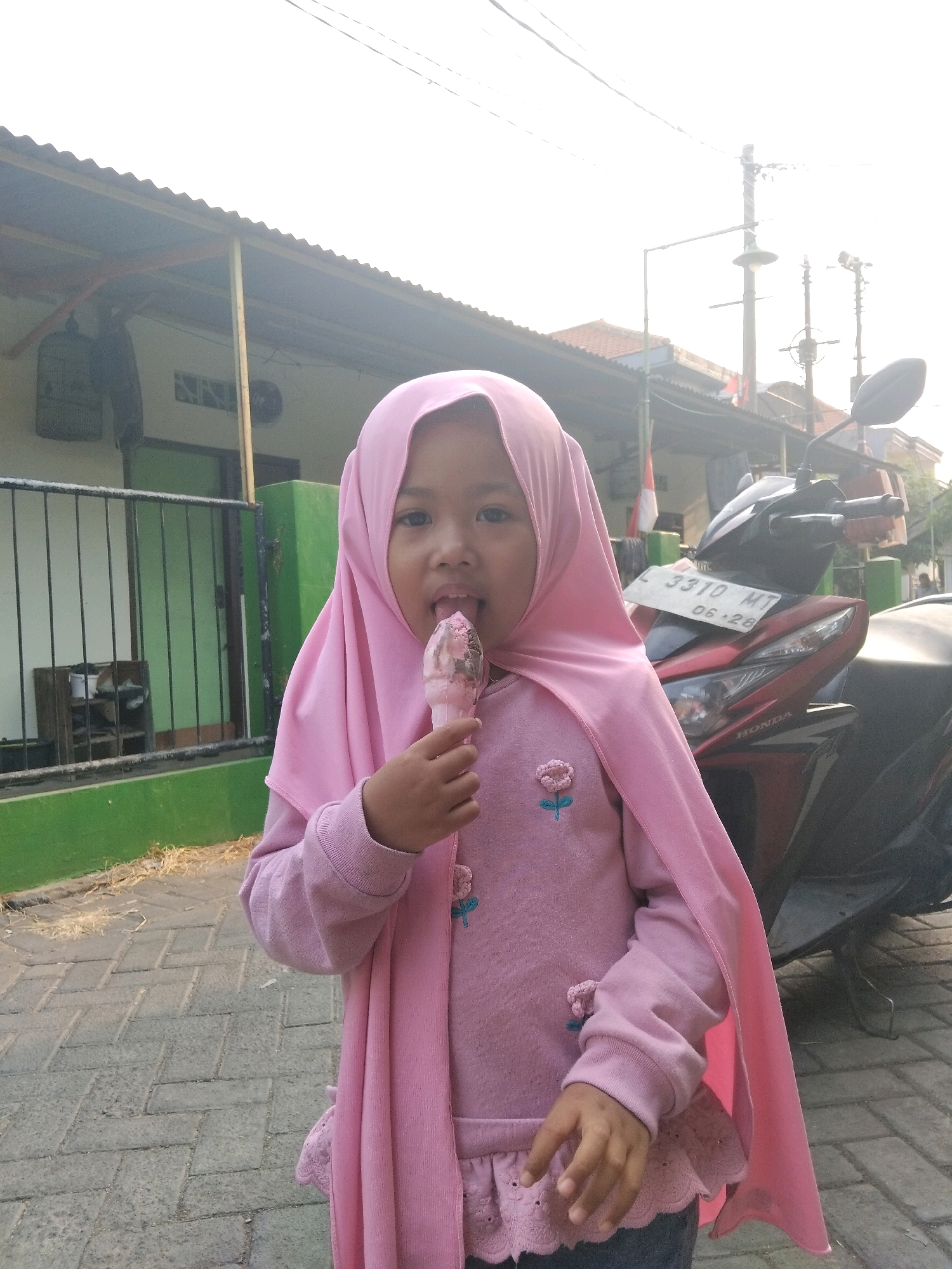 In the city of Kepuh Kiriman, Indonesia, a young girl wearing a pink hijab and pink sweater with flower appliques is captured mid-lick of an ice cream cone. She stands on a brick-paved sidewalk in front of a building with a green wall and corrugated metal roof. A black and red motorcycle with license plate "L 3310 MT" is parked behind her. The background includes a fence, a bird cage hanging from the building, and utility poles with wires, all under a bright, hazy sky.