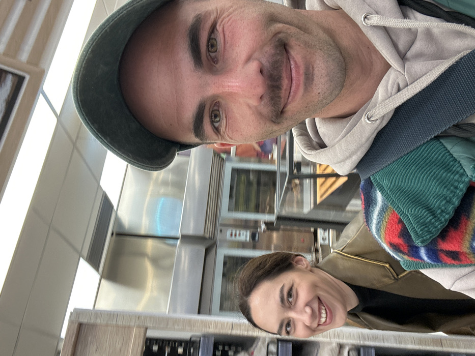 The image depicts two individuals inside an establishment within Budva, Montenegro, appearing to be a bakery or a supermarket's in-store bakery section.
In the foreground, a male subject with light skin, dark hair, and a prominent mustache wears a dark green baseball cap, a light grey hooded sweatshirt, and a multi-colored patterned vest over a dark green corduroy layer. A dark strap crosses his shoulder. He is smiling slightly, looking directly towards the camera, suggesting he is holding the device or posing for a close-up photograph.
Positioned behind and to the left of the male subject is a female subject with light skin and dark hair pulled back. She wears a black top and a brown, leather-like jacket. She is smiling broadly and looking towards the left side of the frame, past the male subject.
The background shows industrial-style stainless steel ovens with glass doors, indicative of a commercial baking area. A clear display case containing items, possibly baked goods, is visible between the two individuals and the ovens. To the far left, shelves with various packaged products are present. The ceiling is white with recessed fluorescent light panels providing illumination to the indoor commercial environment.