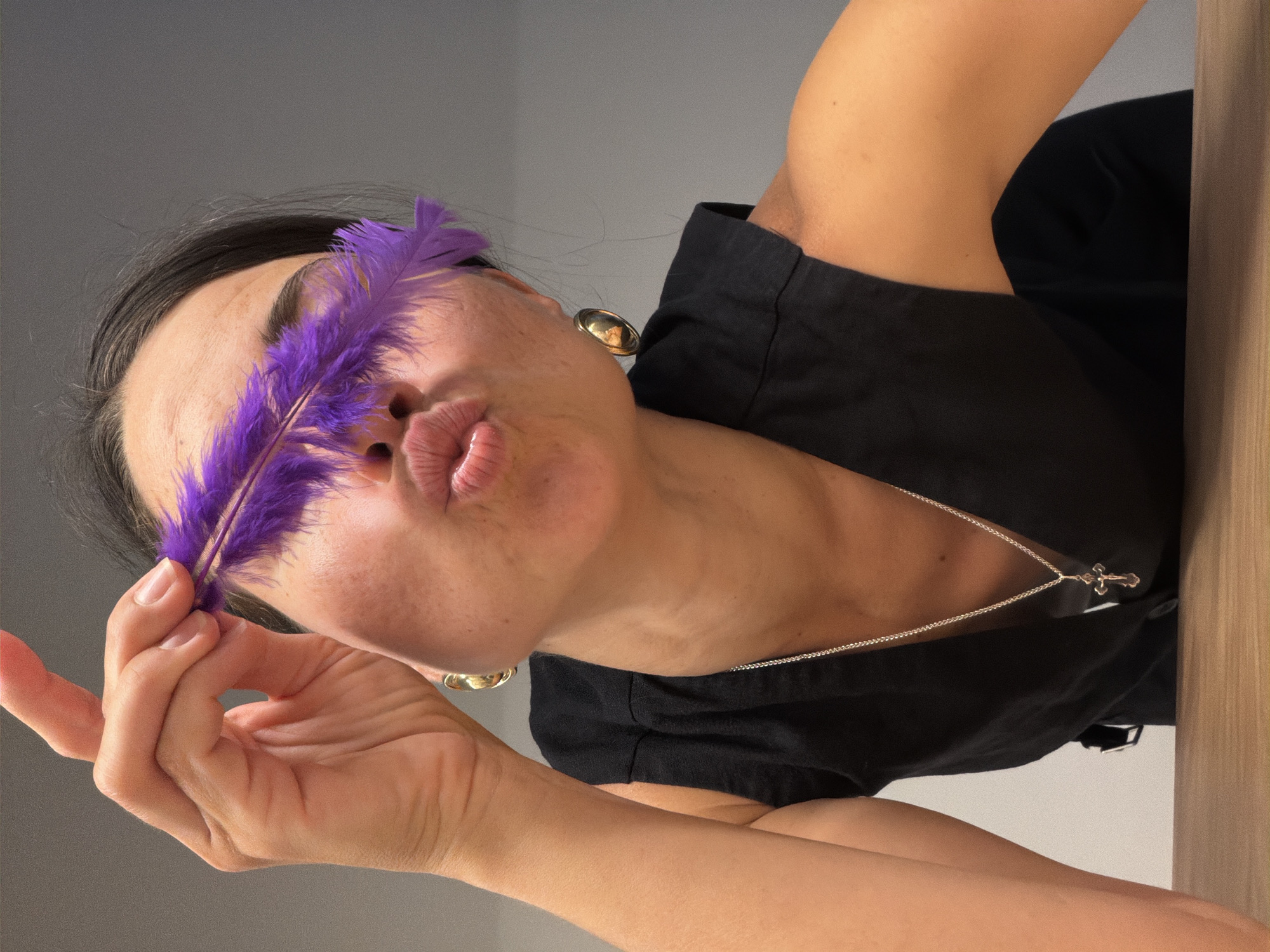 An indoor portrait taken in Budva, Montenegro, features a woman playfully posing with a purple feather. The woman, whose head, shoulders, and arms are visible, holds a vibrant purple feather horizontally across her eyes, obscuring them. Her lips are pursed in a kissing gesture. She has dark hair and is wearing a black top or dress, accessorized with a gold-colored earring and a silver chain necklace with a small cross pendant. The background is a plain grey wall on the left and a light wooden surface on the right. Her hands are visible holding the feather. The overall mood conveyed is playful and lighthearted. No text is visible in the image.
