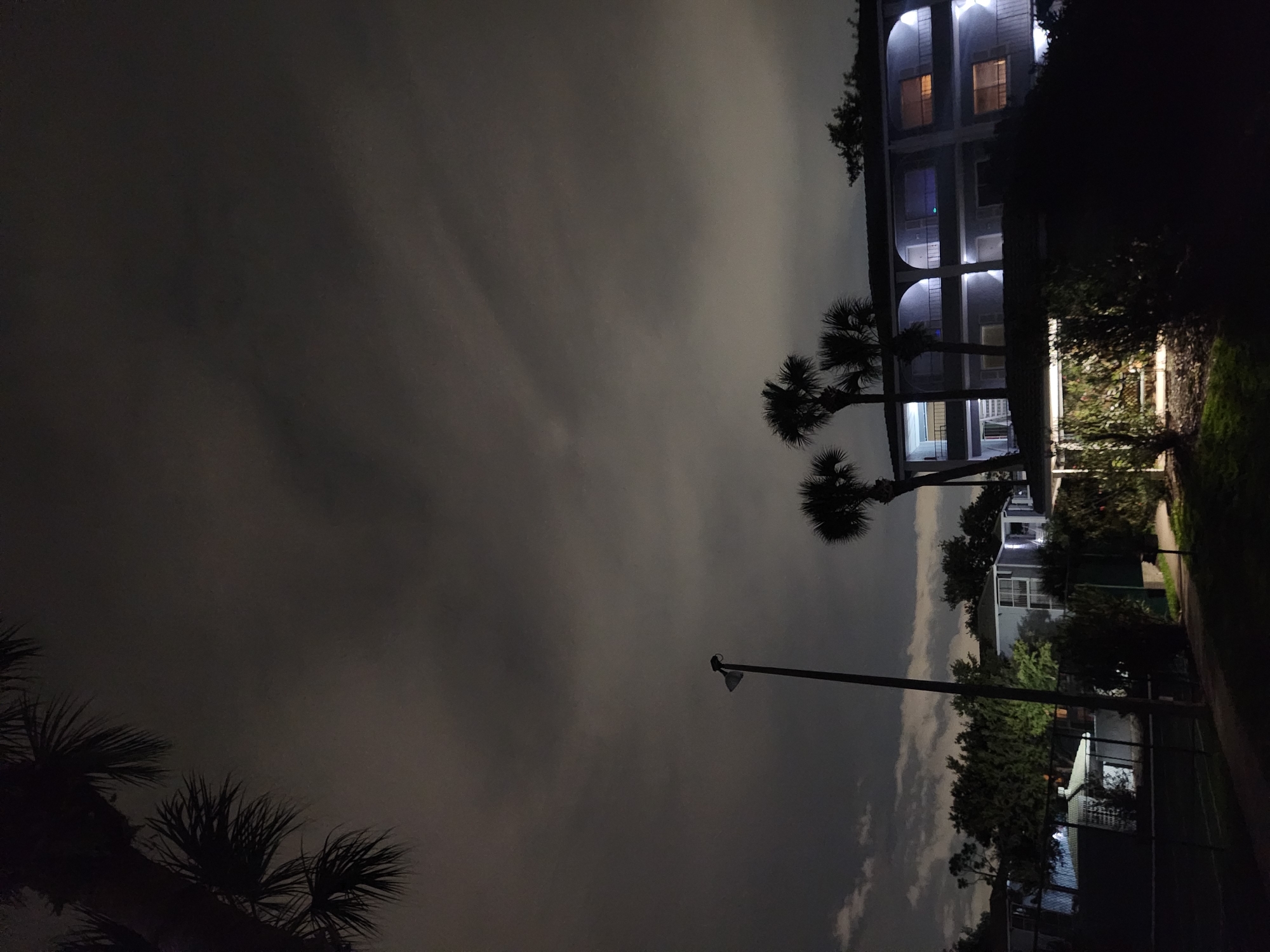 The image depicts an exterior nighttime scene in Altamonte Springs, United States of America. A multi-story building with arched balconies and illuminated windows is visible, surrounded by palm trees and dense foliage. A tall lamppost stands to the left of the building, and a chain-link fence encloses an area in the foreground, which appears to be a sports court. The sky is dark and overcast, with some clouds visible. The lighting is artificial, coming from the building's interior and exterior lights, as well as the lamppost. There are no people or animals visible in the frame.