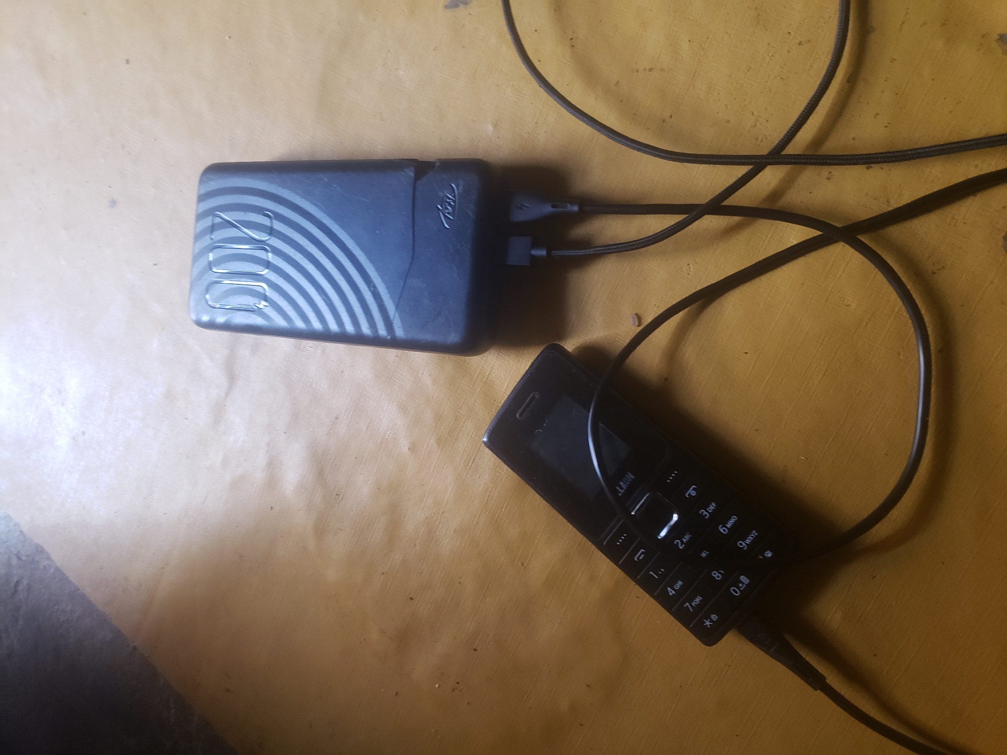 A black feature phone is lying on its side on a wooden surface and is plugged into a dark blue power bank with a black charging cable. The power bank has a circular design on its surface and the brand name "itel" is visible. The phone displays the brand name "LAUN" and has a keypad with numbers and symbols. The wooden surface appears to be a table or floor with some dust particles. The background is partially visible and shows a dark, possibly shadowed area. The scene is located in Rigasa, Nigeria.