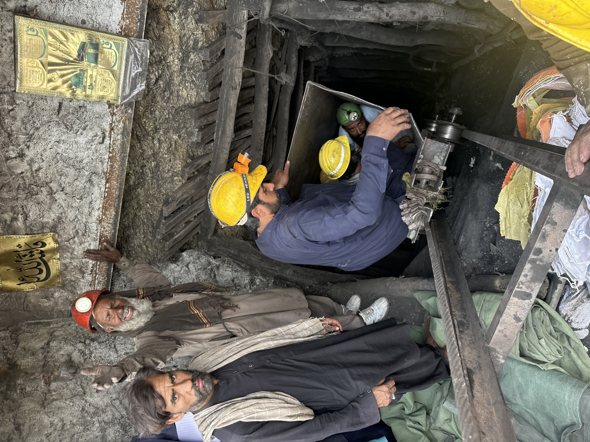 This image depicts a group of men, likely coal miners, at the entrance of a mine shaft in Quetta, Pakistan. In the foreground, a man wearing a black shalwar kameez and a beige scarf is looking directly at the camera with a serious expression. Behind him, an older man with a long white beard, wearing a brown shalwar kameez and a red hard hat with a headlamp, is gesturing with his hand and smiling broadly. He appears to be raising his right hand as if to greet or acknowledge someone. To the right, in the dark mine shaft, two men are visible. One man, wearing a blue shalwar kameez and a yellow hard hat with a headlamp, is leaning into the shaft, holding a metal container. Inside the container, another man is wearing a green hard hat and a headlamp. The mine shaft walls are rough and uneven, reinforced with wooden beams. In the background, a religious tapestry with Arabic calligraphy and an image of the Kaaba is hanging on the wall. The ground is covered with green and yellow bags, possibly containing coal. A metal pulley system is also visible in the foreground, suggesting the method of transporting materials in and out of the mine. The overall scene suggests a working coal mine environment.