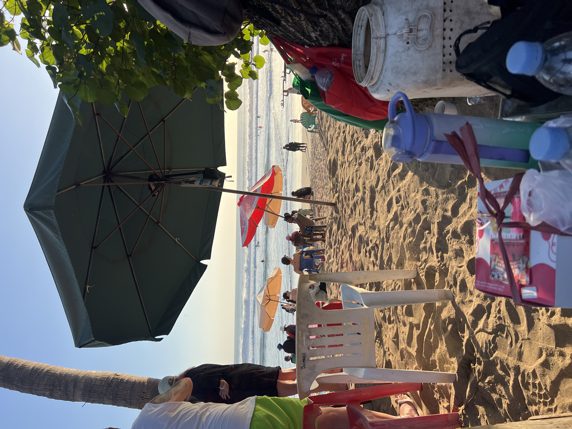 The image depicts a crowded beach scene in Kuta, Indonesia, likely during late afternoon given the warm, golden light and long shadows. The foreground is dominated by sandy ground with various objects scattered on it, including a white plastic chair with visible graffiti, red plastic chairs, a large white container with a lid and a handle, and several colorful bags. A water bottle with a blue and green gradient is also prominent. In the middle ground, people are seated on chairs under umbrellas, facing the ocean. These umbrellas are of different colors, including orange and red. Further out, the ocean is visible with gentle waves and several people surfing or swimming. The background shows a clear sky with a subtle haze near the horizon. A palm tree is visible on the left side of the frame, casting a shadow. The overall atmosphere is one of leisure and recreation.