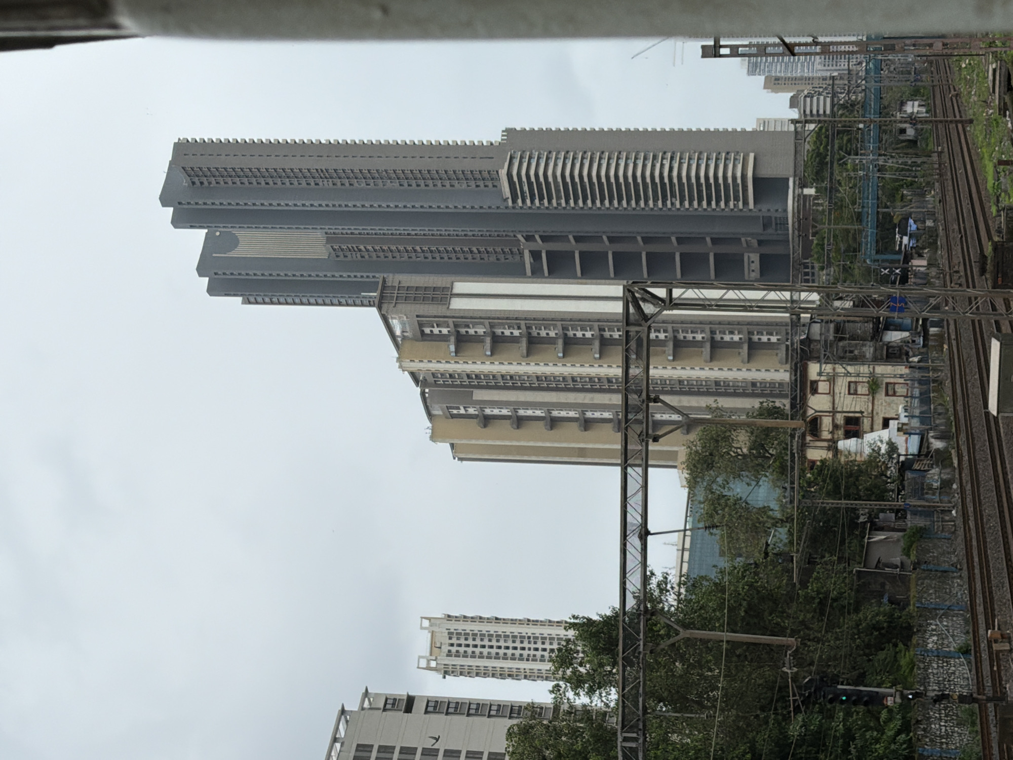 This image depicts a view of Mumbai, India, focusing on a dense urban landscape dominated by modern skyscrapers and railway infrastructure. In the foreground, multiple sets of train tracks run horizontally across the frame, indicating an active railway line. Overhead, a complex network of electrical wires and support structures for the railway system is visible.
Dominating the midground and background are several tall, contemporary buildings. The most prominent is a very tall, gray skyscraper with a distinctive stepped design on its right side and a fluted vertical element on its front. To its left, a cluster of other modern high-rises of varying heights and architectural styles are visible. One building, to the far left, is a light-colored, rectangular high-rise with rows of windows. Further to the right, a lighter-colored, slender skyscraper with many windows stands out. The sky is overcast, creating a muted light across the scene.
Below the skyscrapers, a mix of older structures, trees, and smaller buildings can be seen, suggesting a layered urban development. Some of these lower structures appear to be residential or commercial, with visible windows and balconies. Dense green foliage from trees is interspersed among the buildings, particularly in the midground. A traffic signal with a green light is visible on the left side of the image. The overall impression is one of a bustling, densely populated city with a stark contrast between older and newer architectural elements.