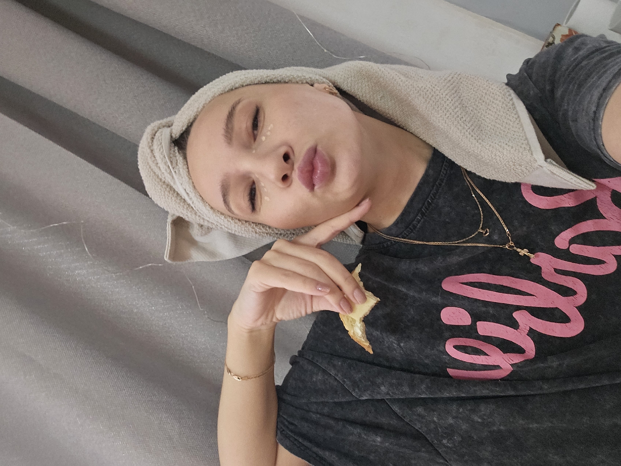A young woman with fair skin and dark eyebrows is captured in a selfie-style portrait. She has a towel wrapped around her head, suggesting she may have recently washed her hair or performed a skincare routine, as evidenced by the small dots of cream on her face, specifically under each eye. Her lips are pursed in a "duck face" pose.
She is wearing a dark gray t-shirt with a large, cursive inscription in bright pink, and a thin gold necklace with a small pendant. On her left wrist, she wears a delicate gold bracelet. Her left hand is near her face, and she is holding a piece of what appears to be a pastry or chip between her fingers.
The background consists of gray curtains or drapes.
While there is no direct indication of the city of Chaikovsky, Russia, in the image, this information was provided, suggesting that the photo was taken in this location.