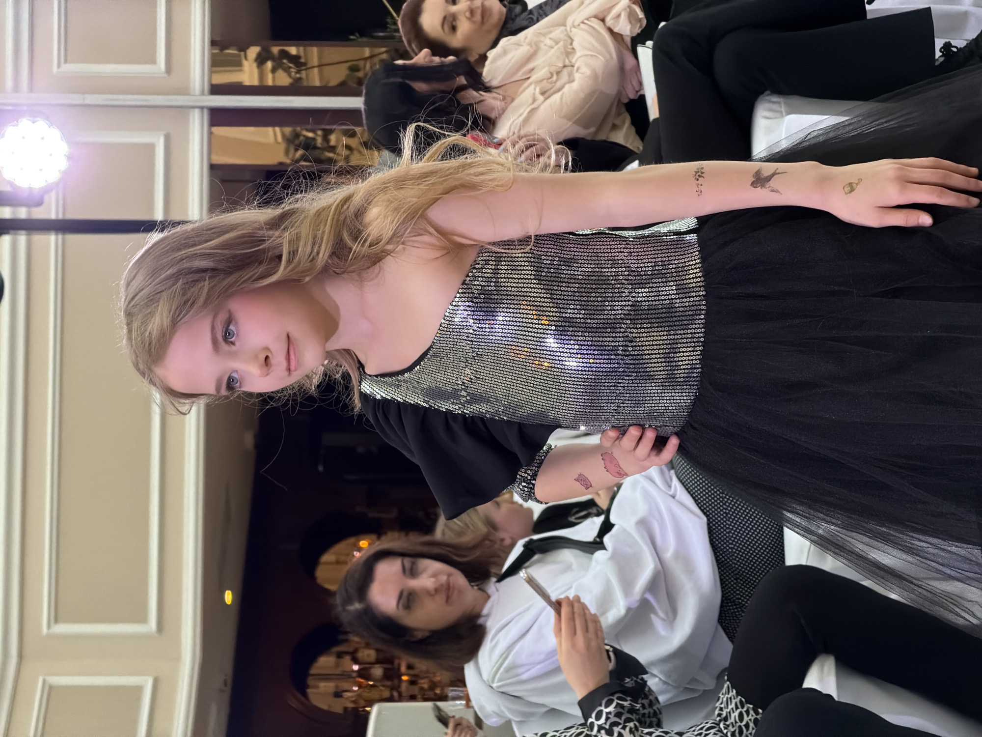 A young, blonde-haired girl in a sequined top and black tulle skirt looks toward the camera. She has temporary tattoos on her arms. She is seated, surrounded by other people, presumably at a fashion show or similar event. Other women are seated around her, some in white shirts. The setting is indoors, possibly a venue in Saint Petersburg, Russia, given the architectural details visible in the background, including arched openings and paneled walls. A stage light is visible to the left.