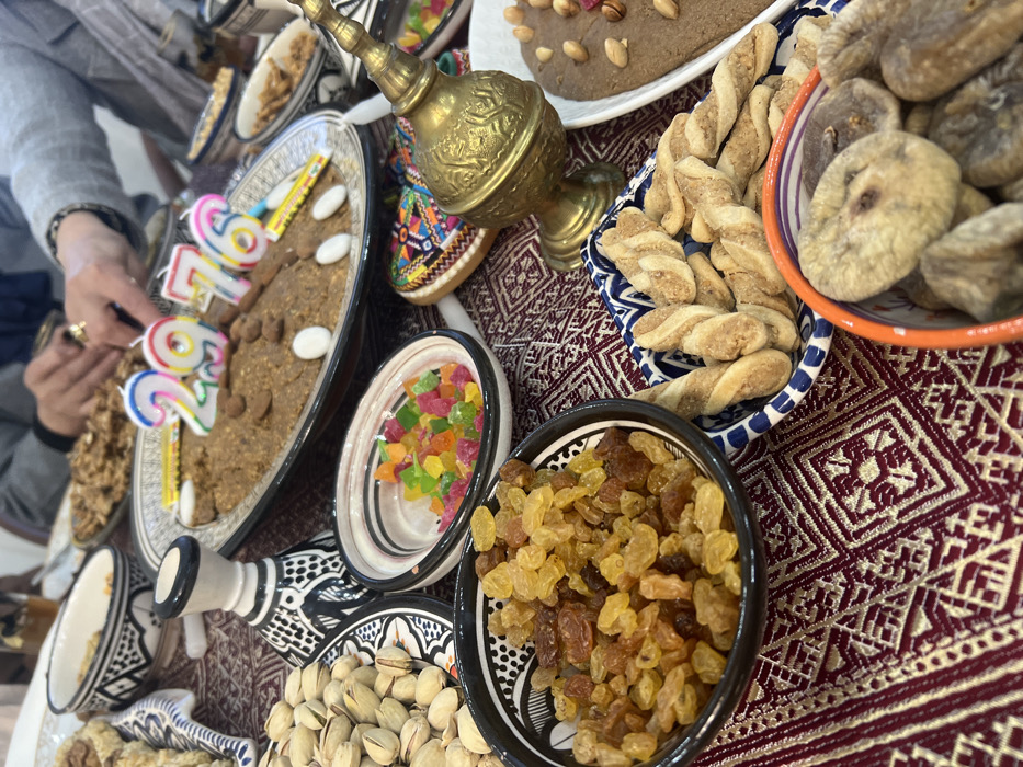 A table displays various foods including a light brown cake with birthday candles spelling out "29", along with almonds and white candy. Surrounding it are bowls of dried figs, raisin, candies, and pistachios. A silver pot sits beside these, while a dark brown cake with almonds decorates another section. Twisted pastries are also visible, and the table is covered with a maroon and white patterned cloth.