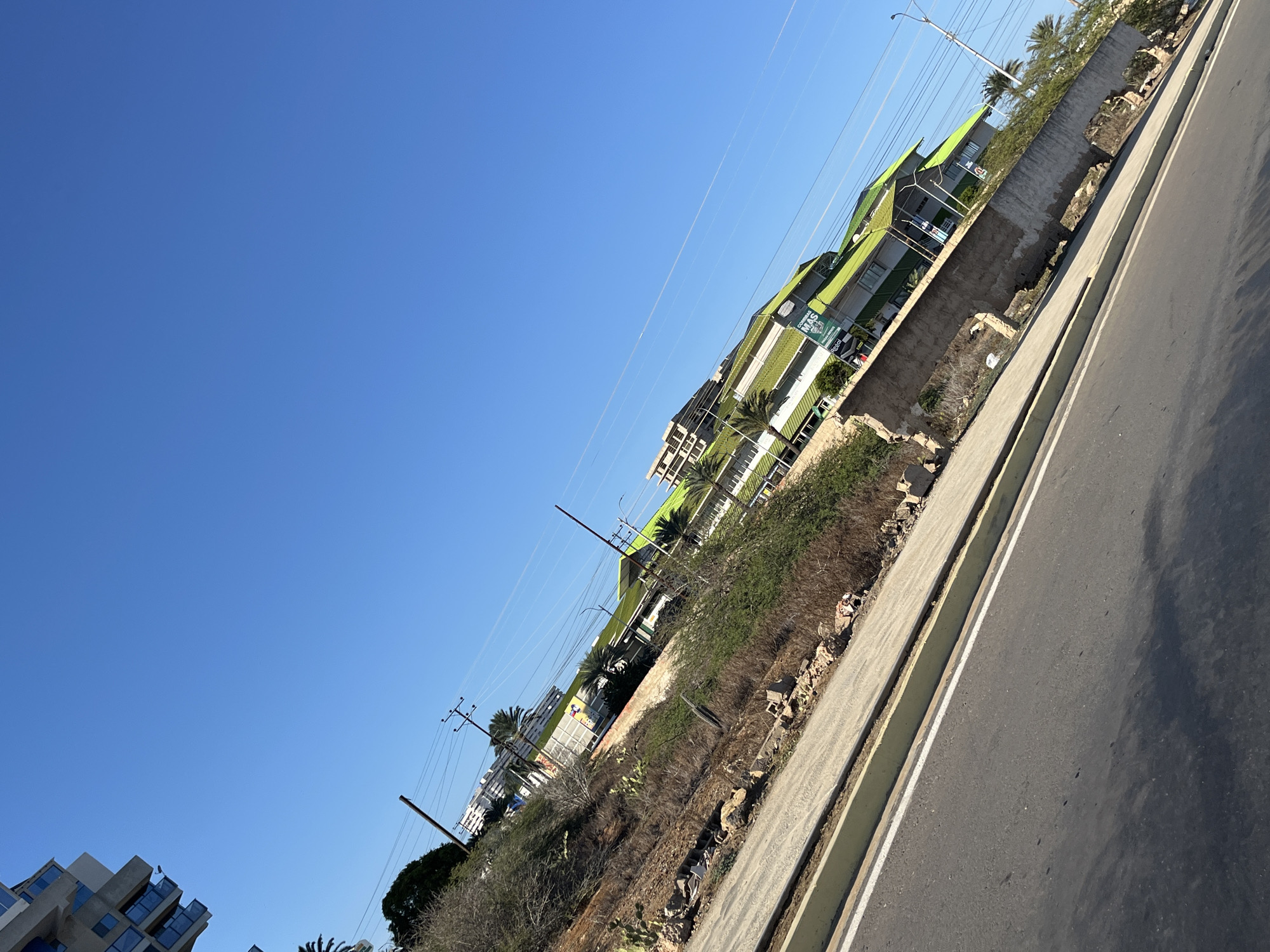 The image captures an urban or suburban landscape in Porlamar, Venezuela, under a brilliant, cloudless blue sky. The photograph is taken from a highly tilted angle, placing the sky prominently in the upper-left two-thirds of the frame, while the ground and structures extend diagonally across the lower-right.
The expansive sky is a vibrant, clear blue, indicative of a sunny day with excellent visibility, likely morning or afternoon. Faint, thin lines, possibly distant contrails or power lines, subtly traverse the sky.
In the lower-right, a paved asphalt road with a distinct white lane marking runs diagonally. Adjacent to the road is a concrete sidewalk or curb. Beyond this, a sloped embankment of dry earth, sparse brush, small cacti, and scattered rocks extends towards a row of buildings. Parts of a crude concrete retaining wall are visible along this slope, appearing somewhat worn or unfinished.
Along the upper-right diagonal, several multi-story buildings feature distinctive bright green roofs or awnings. These structures, potentially residential or commercial, display numerous windows and balconies. One building clearly exhibits a white sign with the Spanish text "COMERCIOS MÁS" and "REALIZACIONES" below it, accompanied by a logo. In the lower-left corner, a portion of another building with an angular, modern design and dark-framed glass windows is visible.
Numerous utility poles, both wooden and metal, supporting a network of overhead power lines, are scattered throughout the scene, particularly towards the lower-left and mid-frame, reaching into the sky. A tall street light pole stands near the upper-right. Palm trees and dense green foliage are visible around and behind the green-roofed buildings, providing a contrast to the drier vegetation on the embankment.
No people or specific activities are depicted, making the image a static portrayal of this bright, sunlit environment.